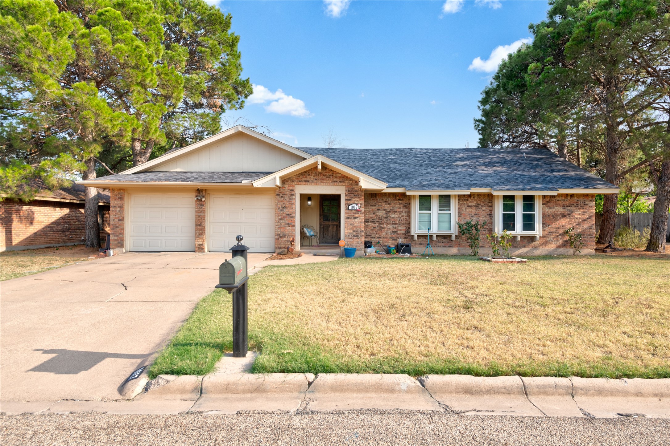 Single-story brick house with a two-car garage, broad driveway, and mature pine trees.