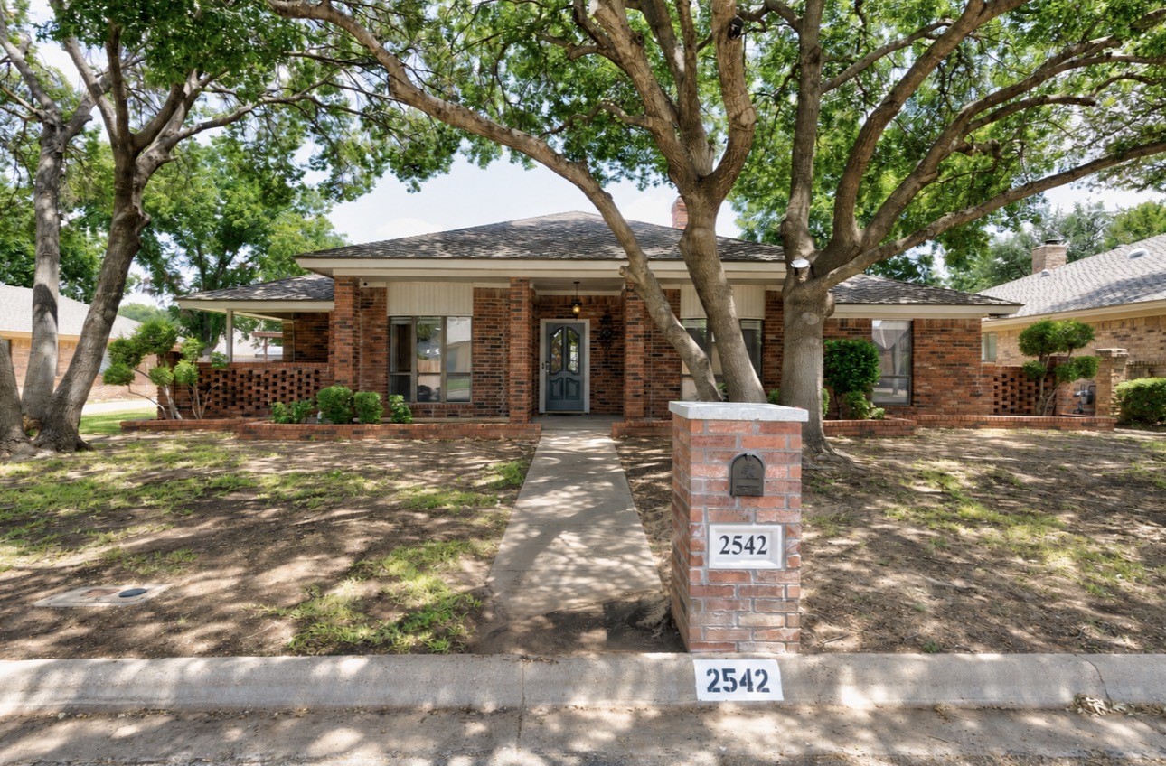 Single-story brick house shaded by mature trees, with a walkway and address 2542 on the mailbox pillar.