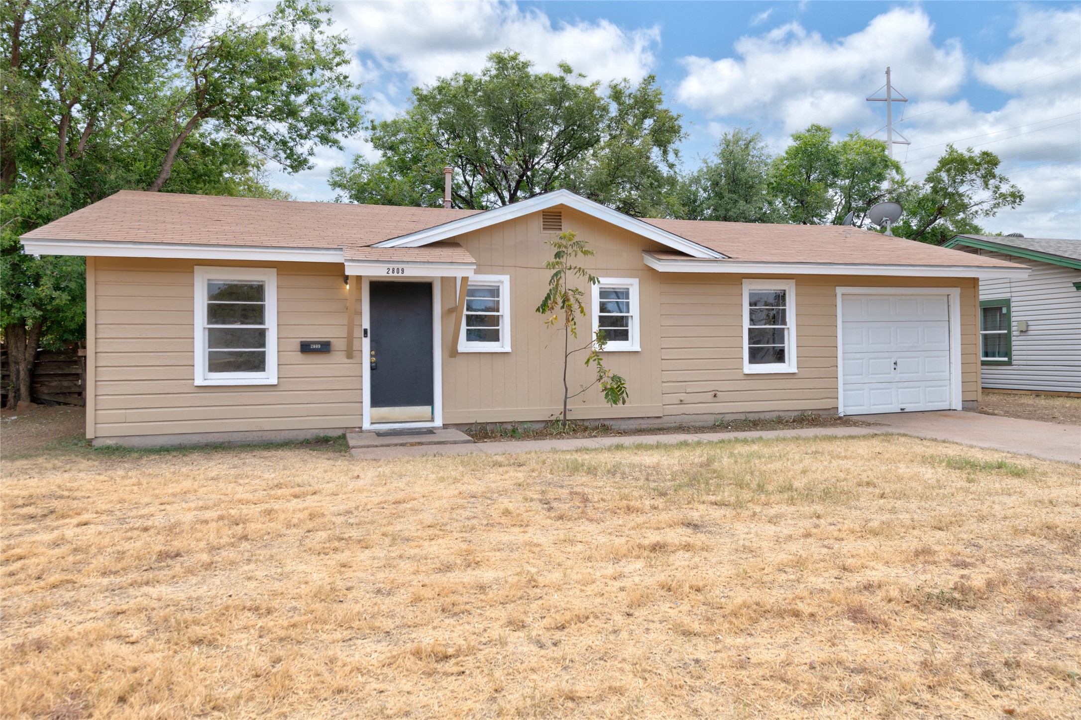 Single-story tan house with an attached garage, dark front door, and dry lawn.