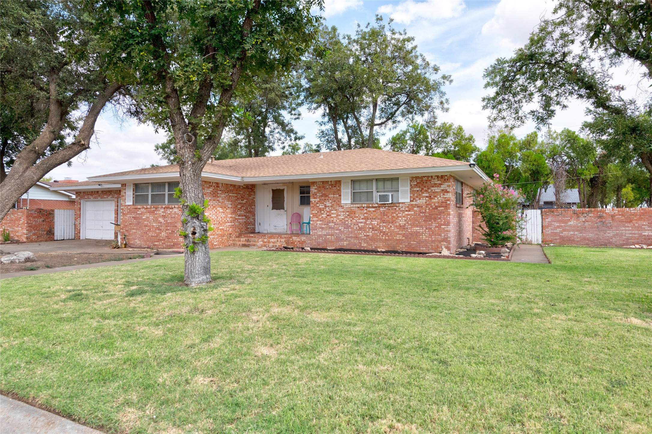 Single-story brick house with a broad lawn, mature shade trees, and a garage.