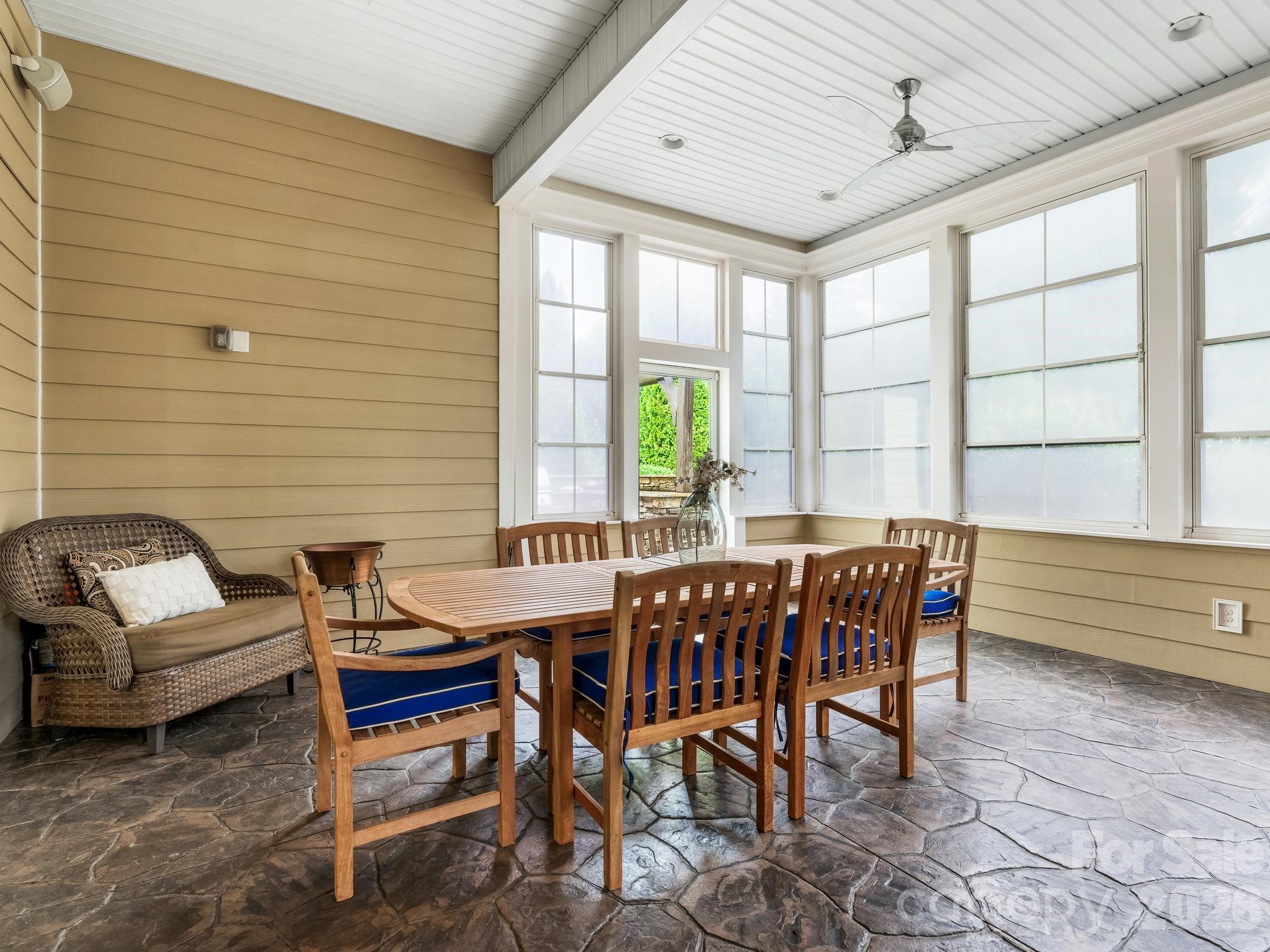 Furnished sunroom with a wooden dining set, wicker lounge chair, and large windows.