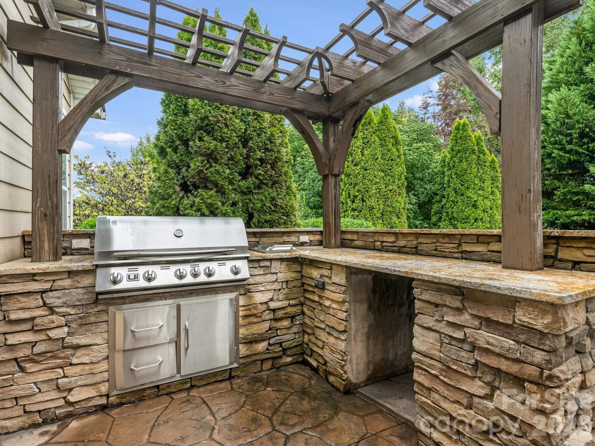 Outdoor kitchen with built-in stainless-steel grill, stone counters, and a wooden pergola.