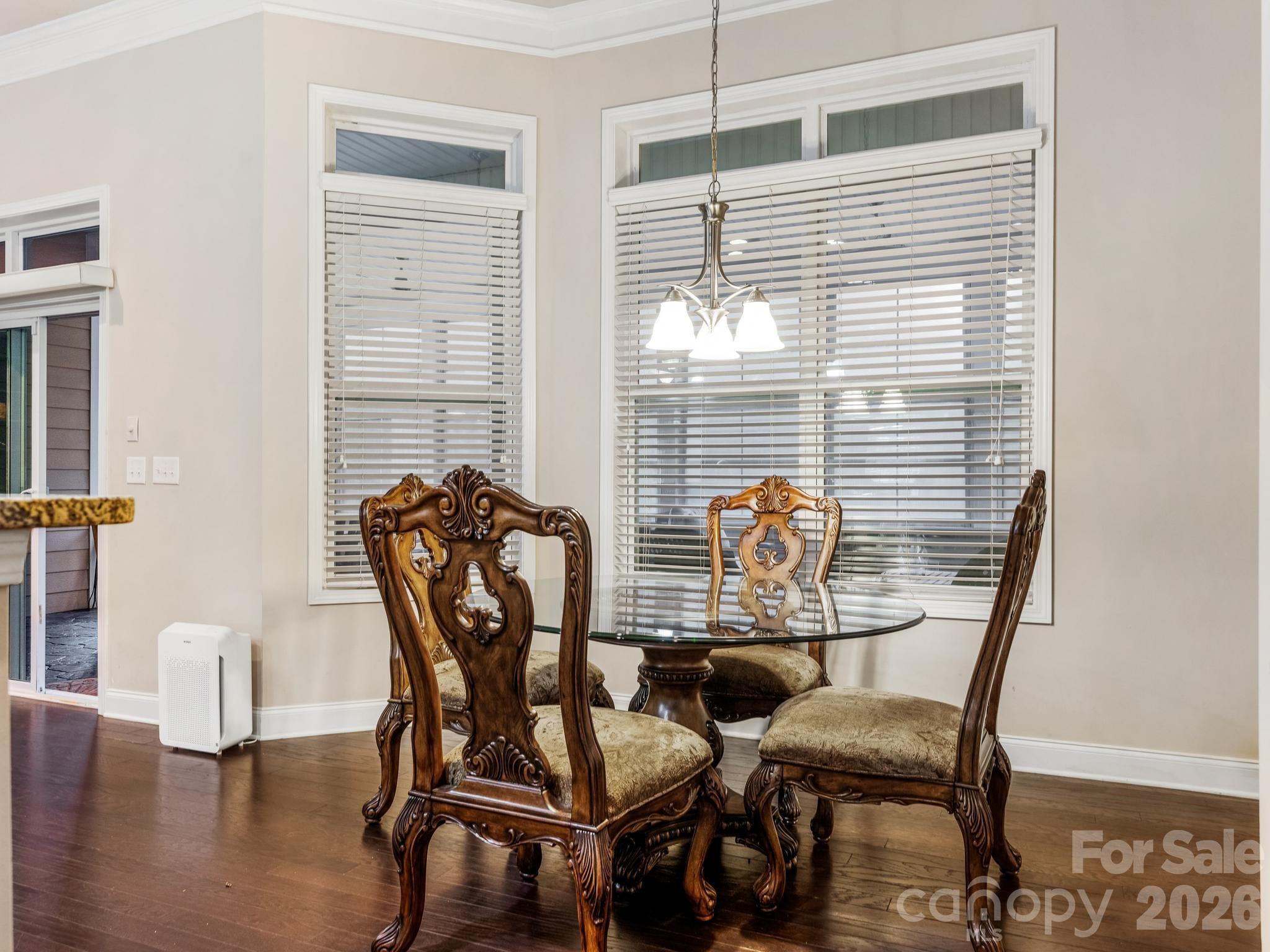Dining room with ornate carved chairs around a glass-top table beneath large windows.