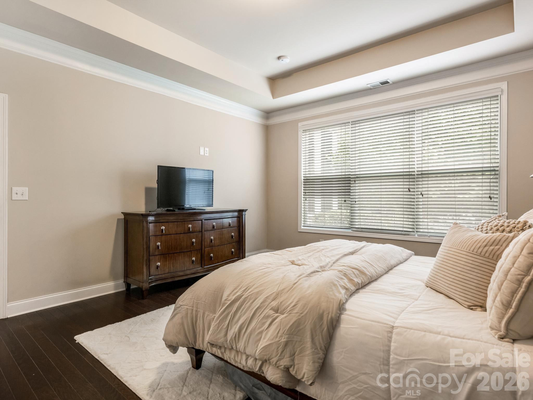 Bedroom with cream bedding, dark wood dresser and television, and a wide window with blinds.