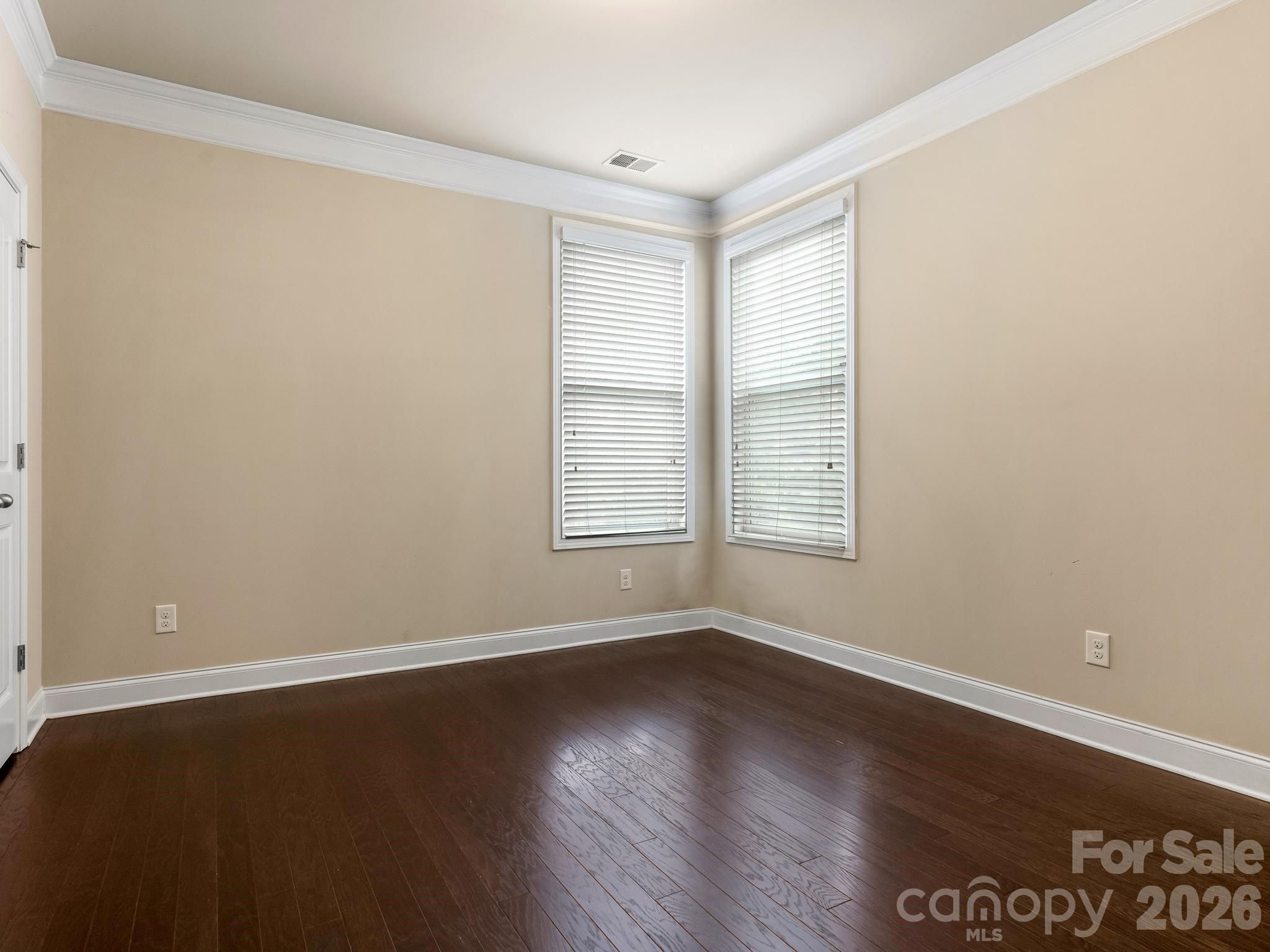Empty room with dark hardwood floors, beige walls, and two windows with white blinds.