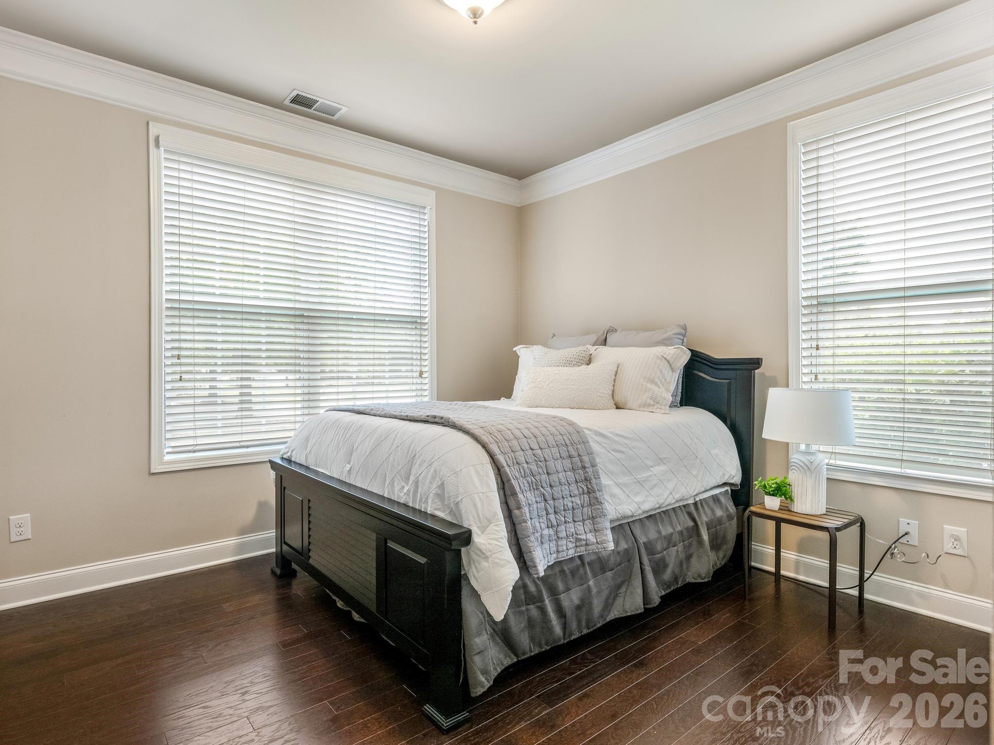 Furnished bedroom with a dark wood bed, layered white and gray bedding, and two large windows.