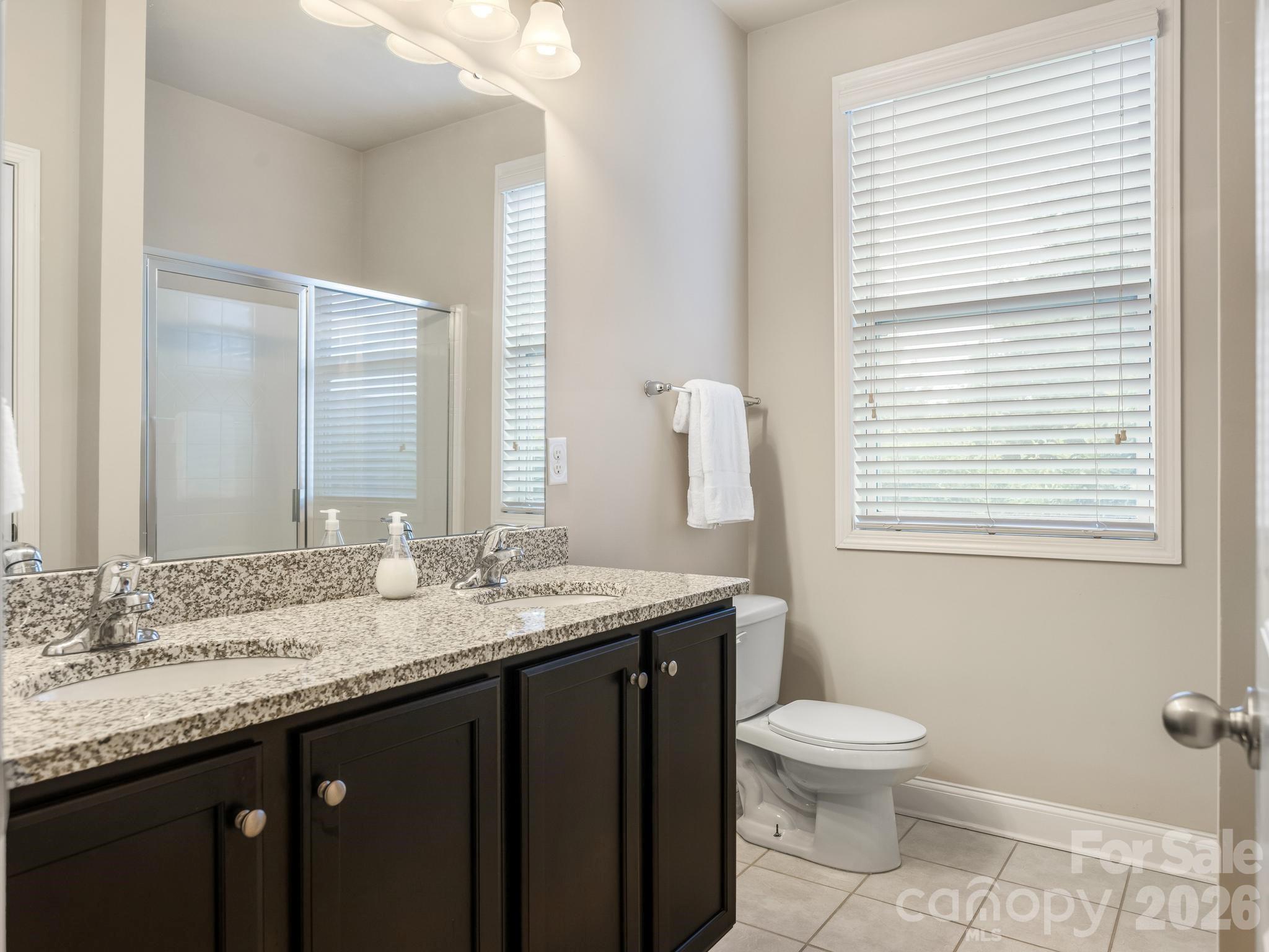 Bathroom with a speckled granite double vanity, glass shower, and large window with blinds.