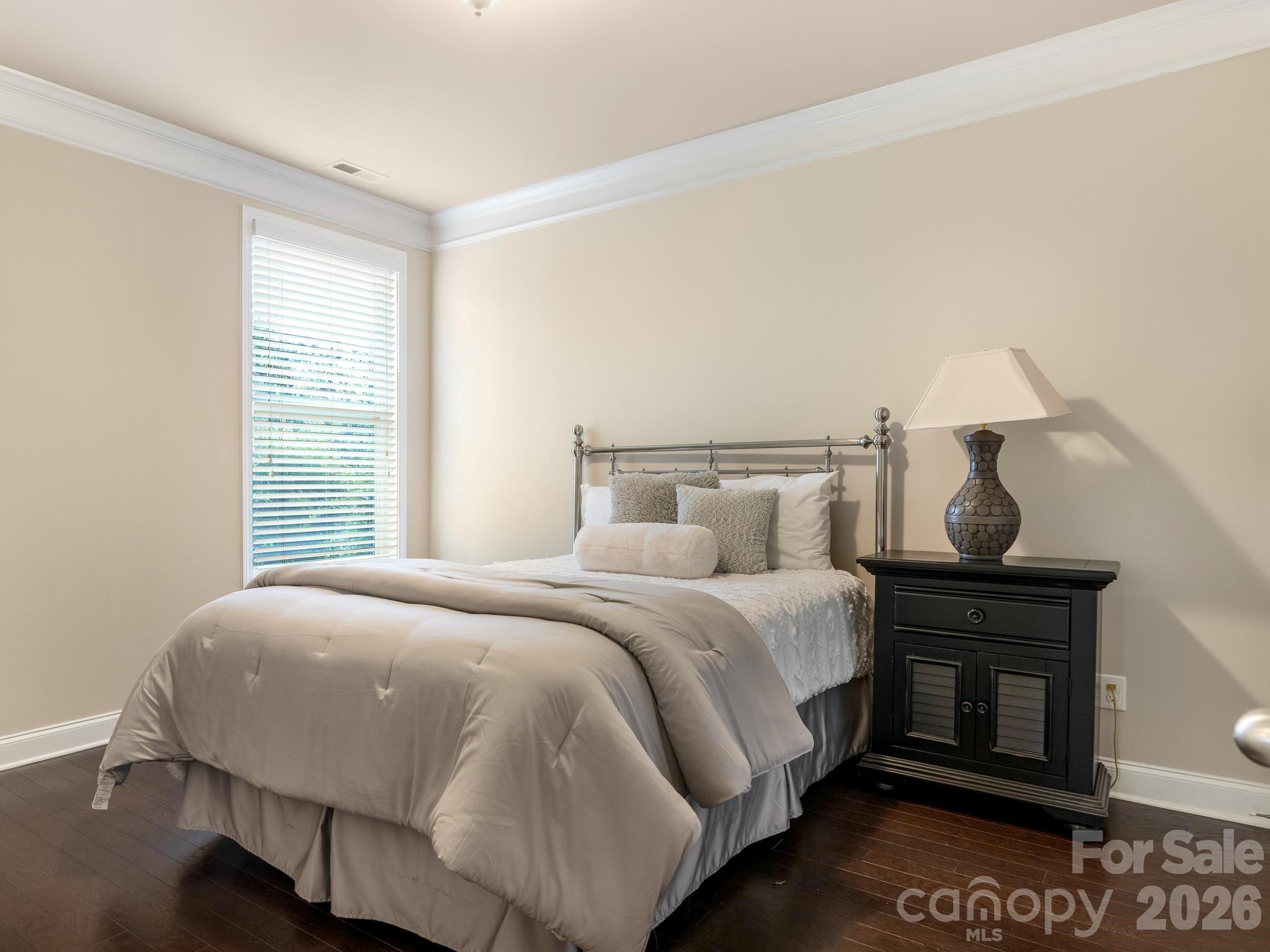 Staged bedroom with gray-and-white bedding, a tall window with blinds, and a black bedside cabinet.