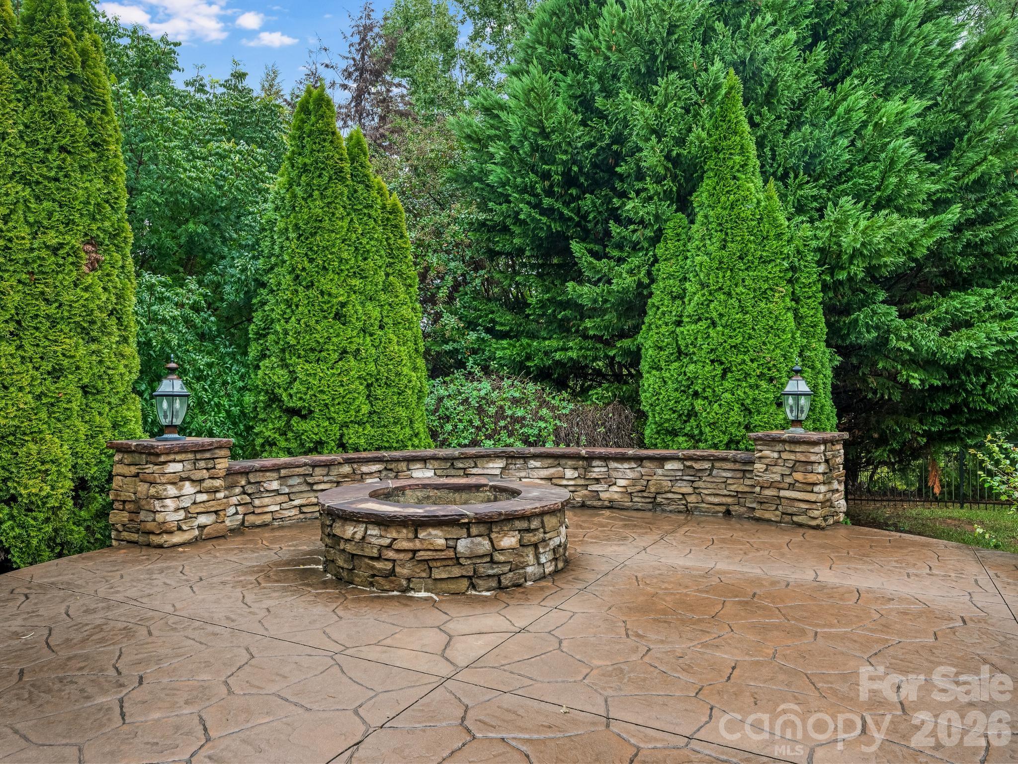 Stone patio with a circular fire pit, low stone wall, and dense evergreen landscaping.
