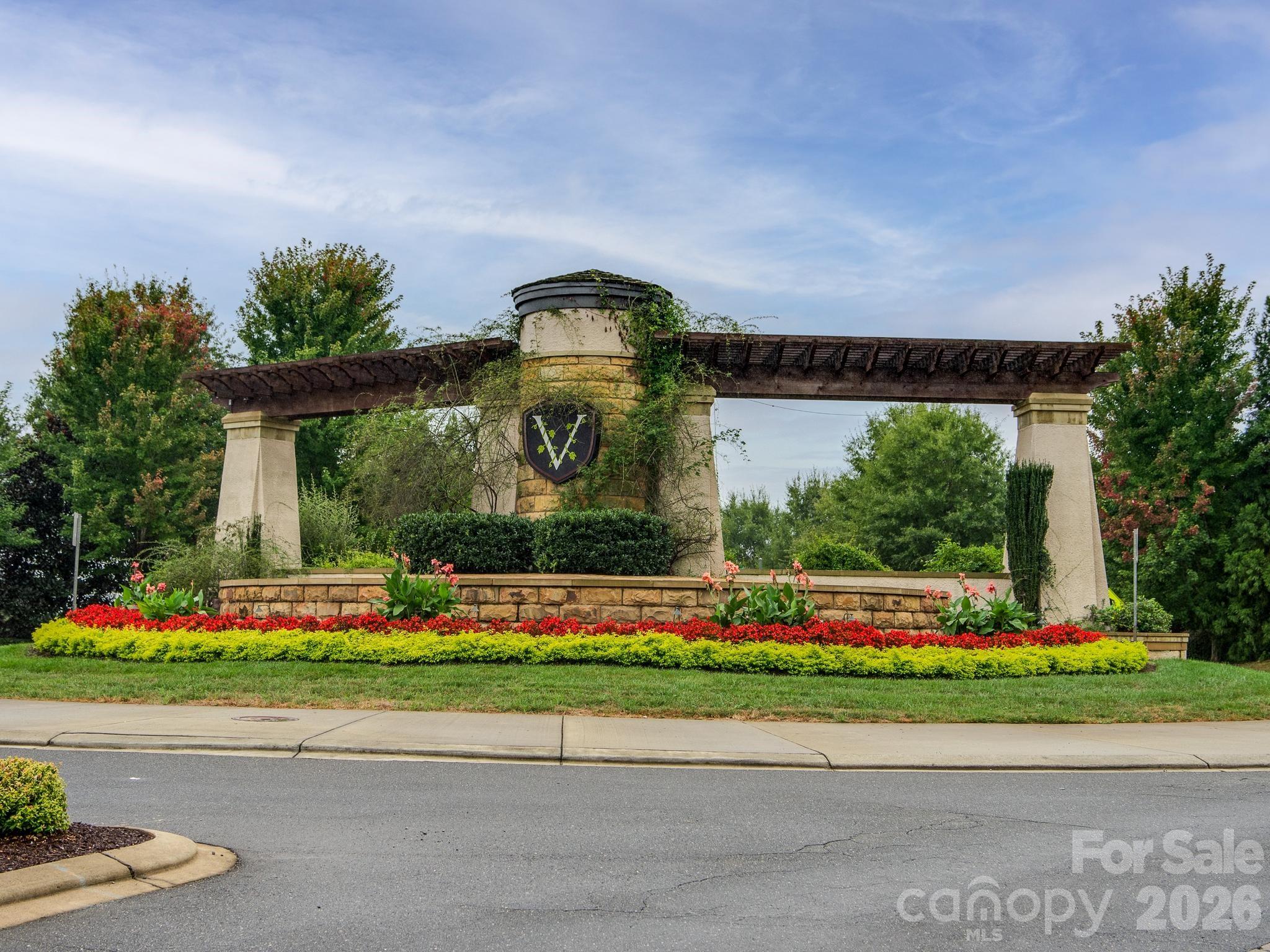 Landscaped neighborhood entrance with a stone pillar, wooden pergola, and colorful flower beds.