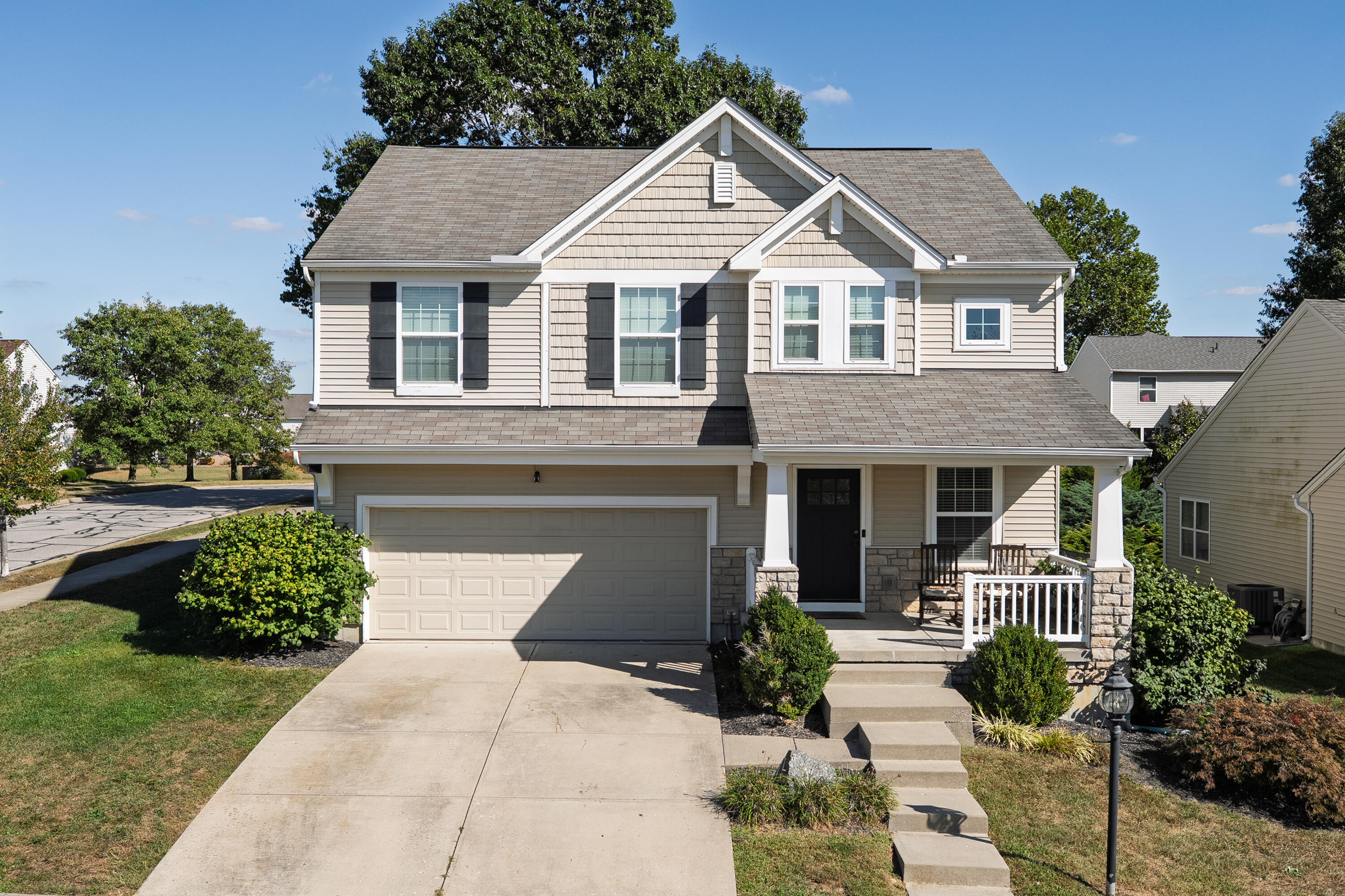 Two-story beige house with a two-car garage, stone porch supports, and white railing.