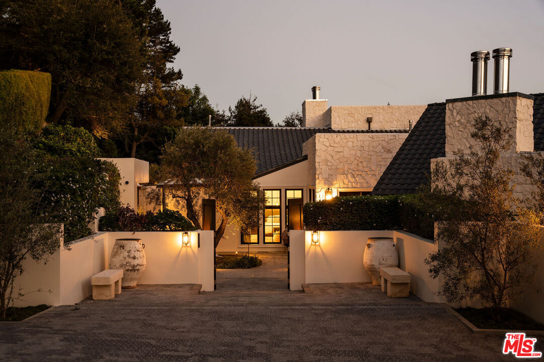 Modern home entrance at dusk, with pale stone walls, dark tiled roofs, and a warmly lit driveway.