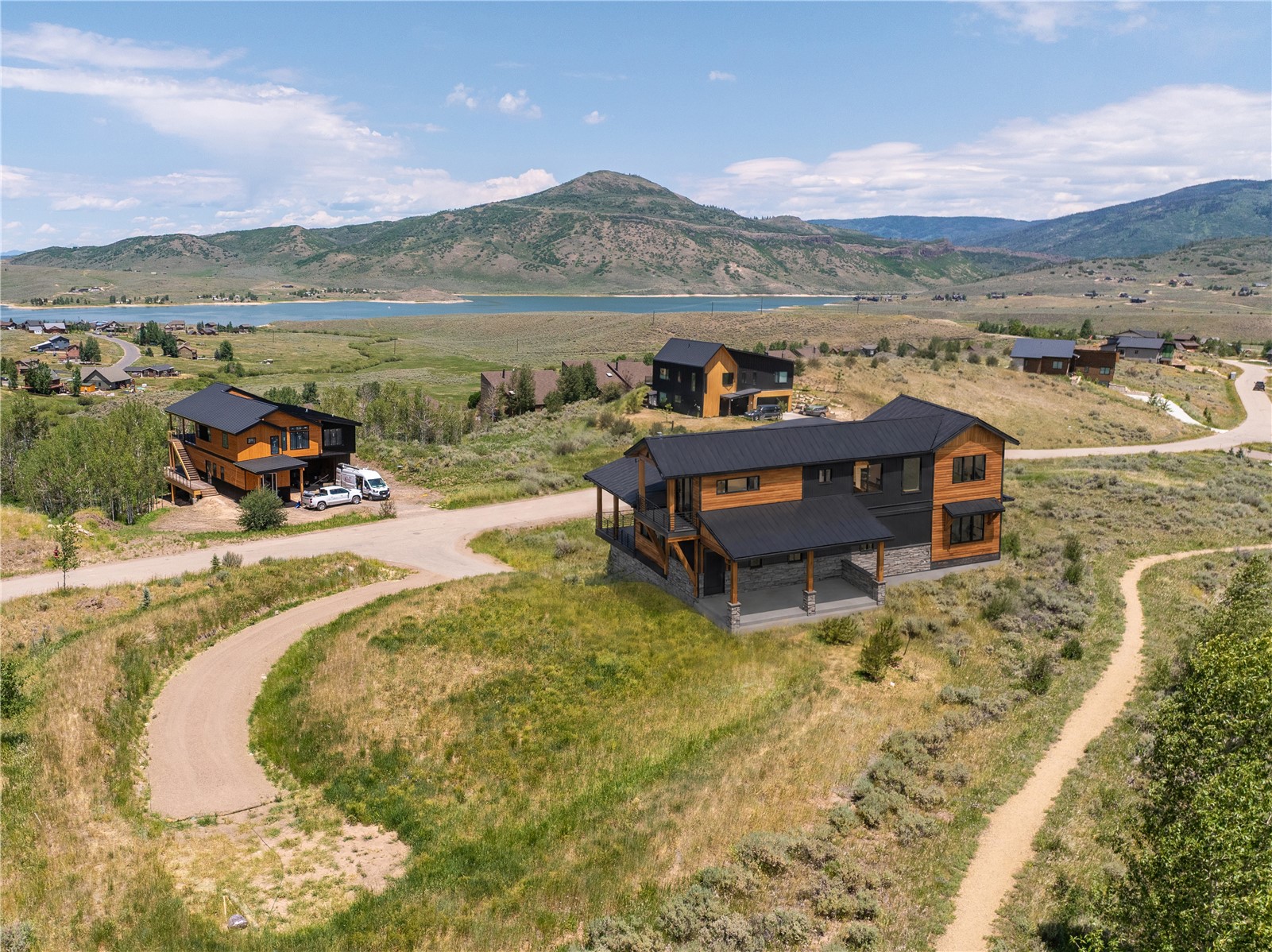 Aerial view of a modern mountain home beside winding roads, lake, and hills.