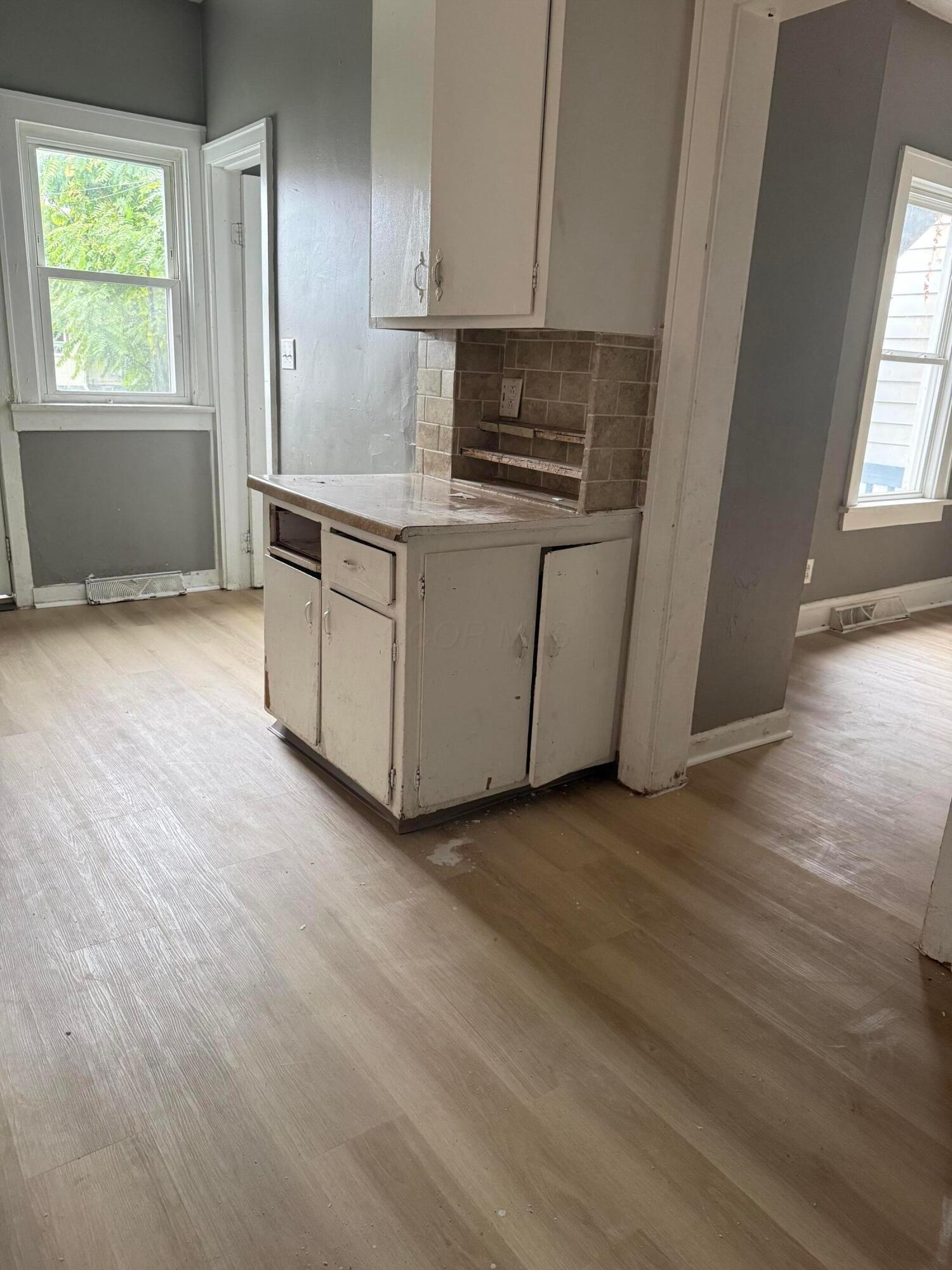 Dated kitchen with worn white cabinets, brown tile backsplash, and windows on two sides.