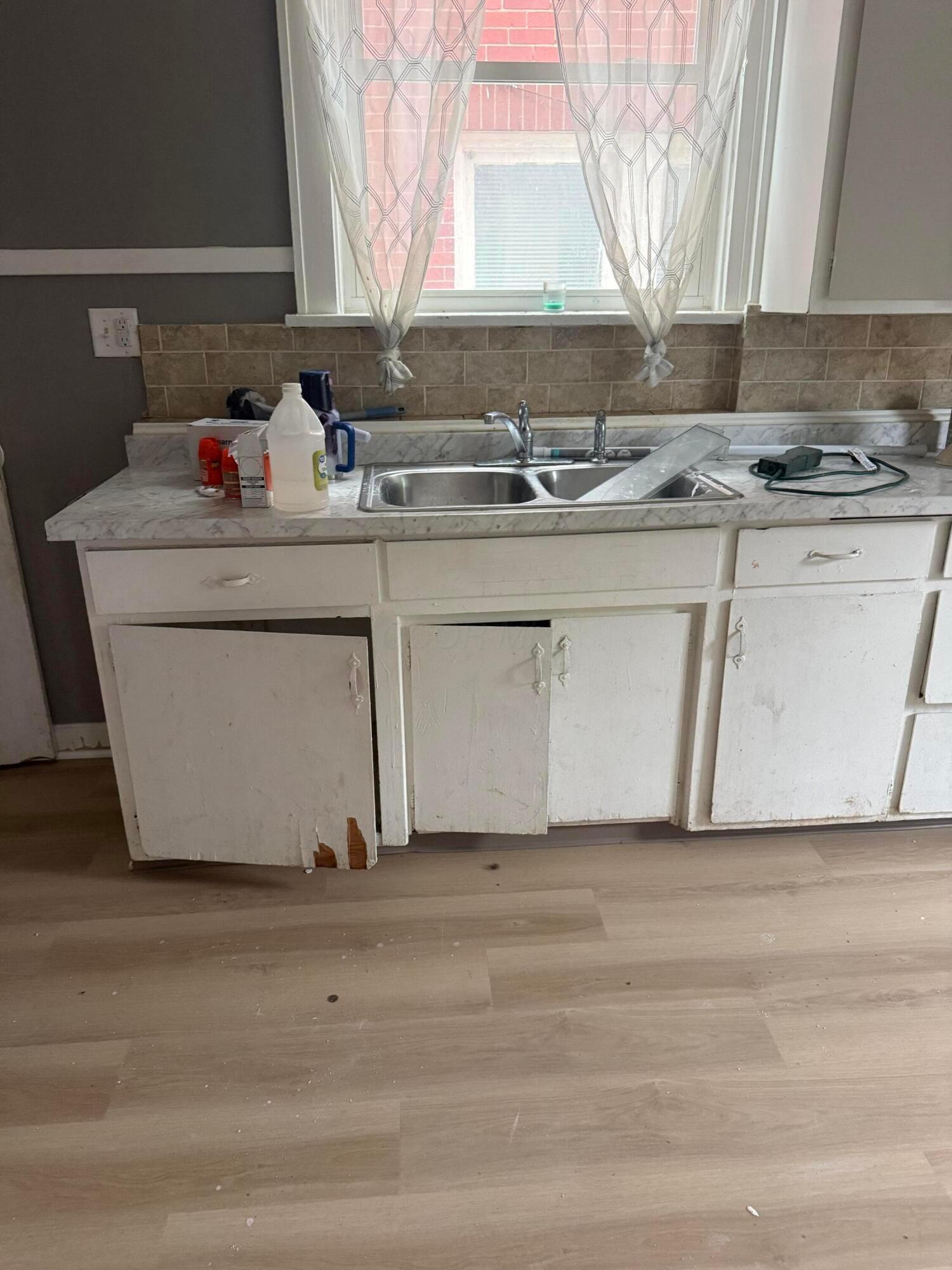 Kitchen sink beneath a window, with a marble-pattern counter and worn white cabinets.