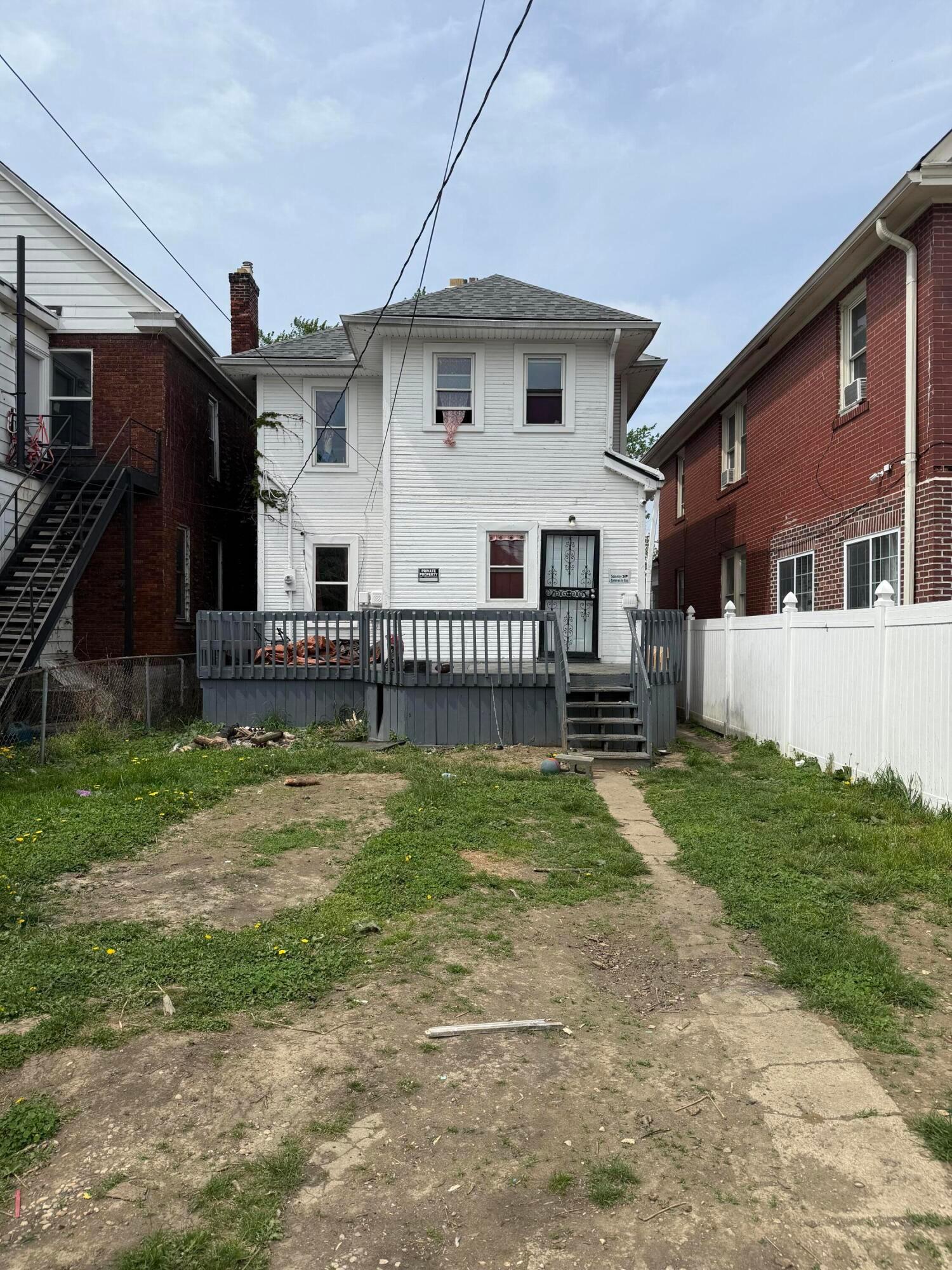 White two-story house with a raised gray deck and patchy backyard between neighboring homes.