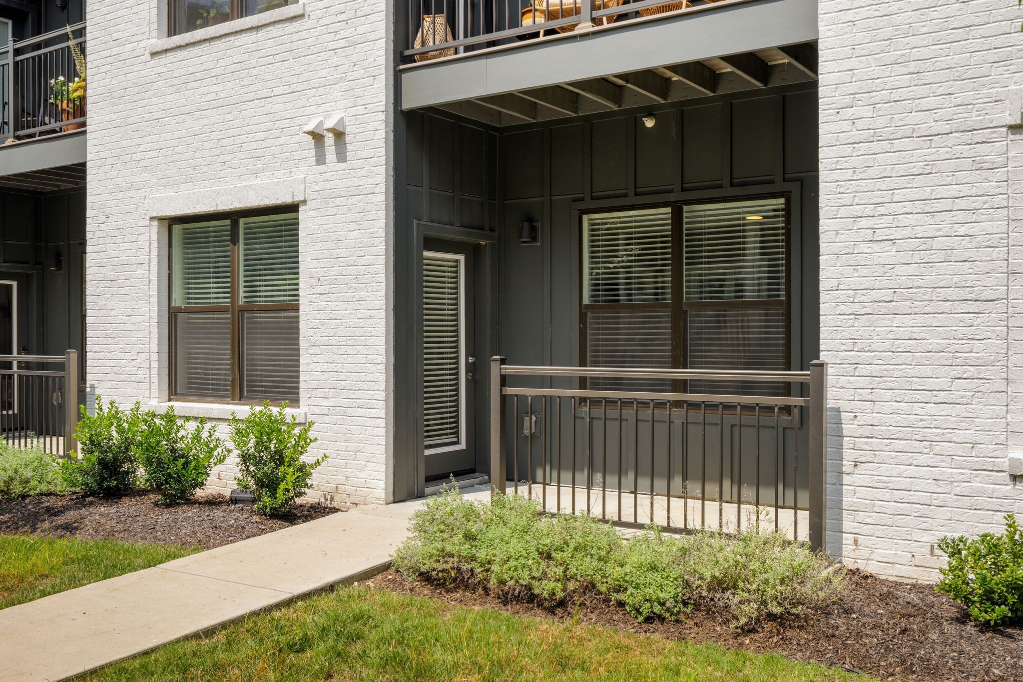 Ground-level condo entrance with white brick, a covered patio, metal railing, and landscaped shrubs.