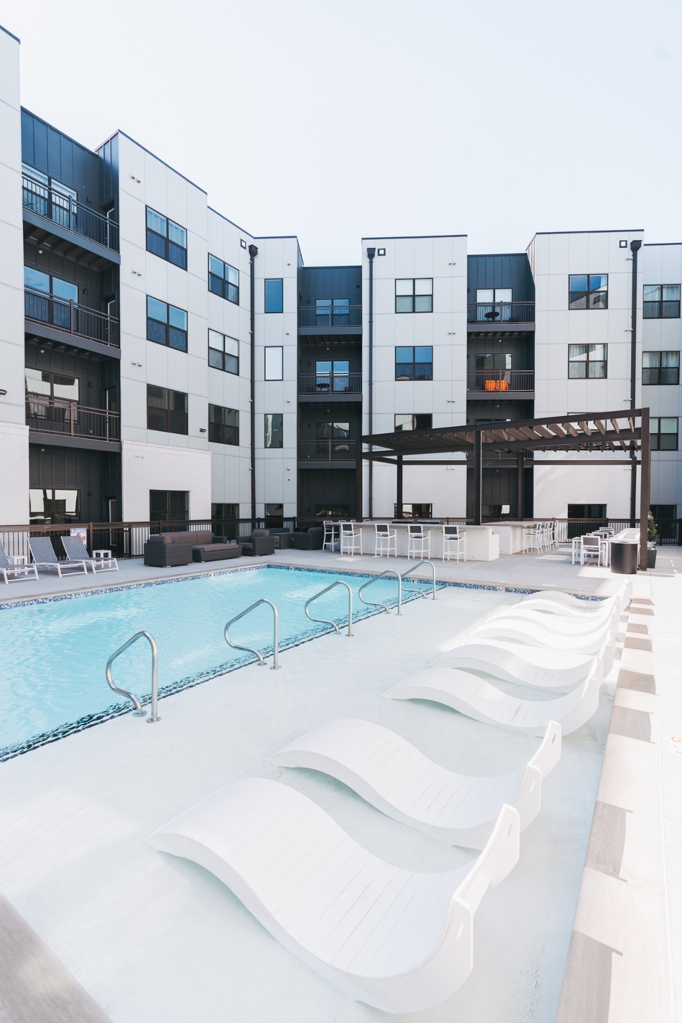 Outdoor pool with white wave-shaped loungers and a pergola beside a modern apartment building.