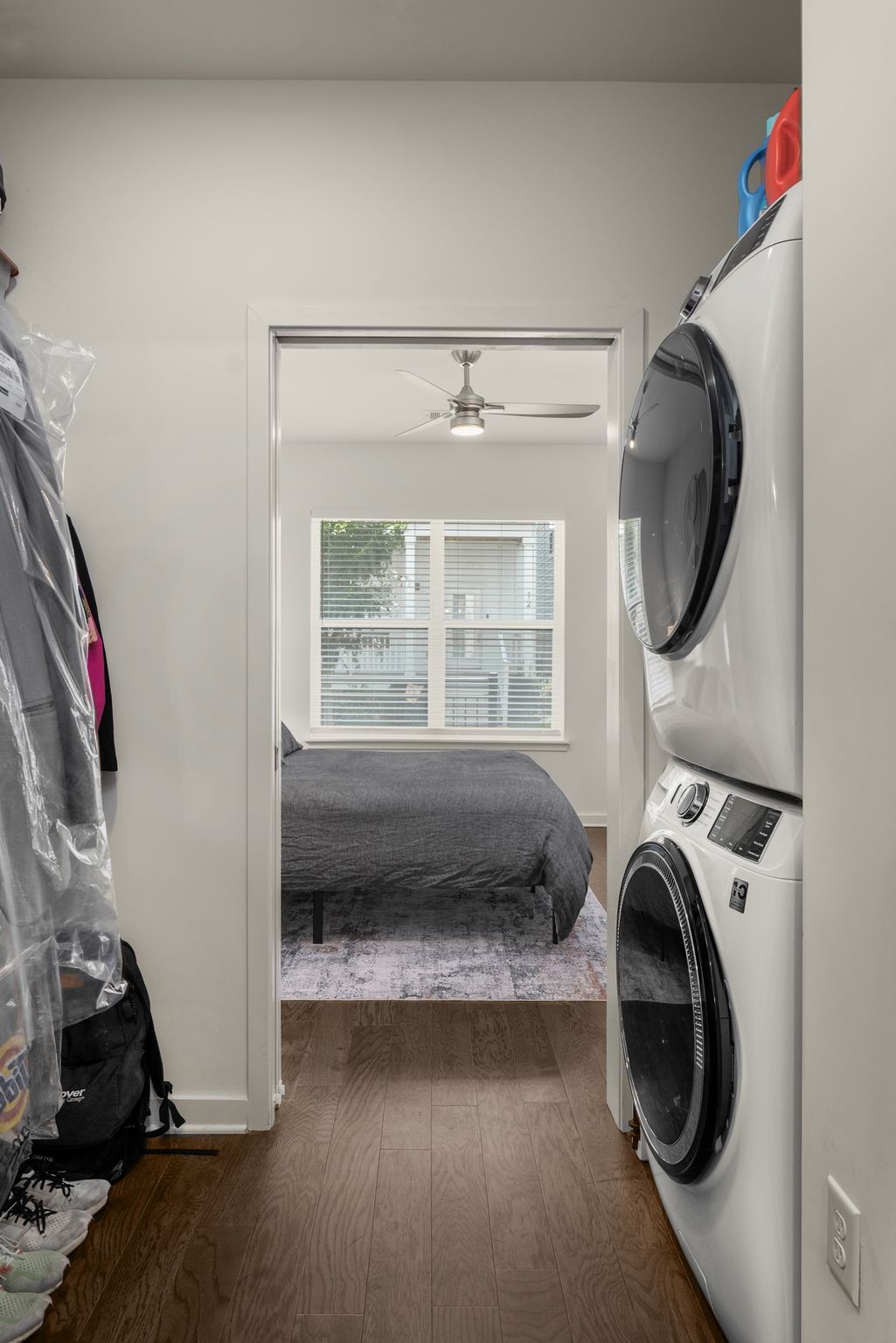 Stacked washer and dryer beside a bedroom with a gray-covered bed and window blinds.