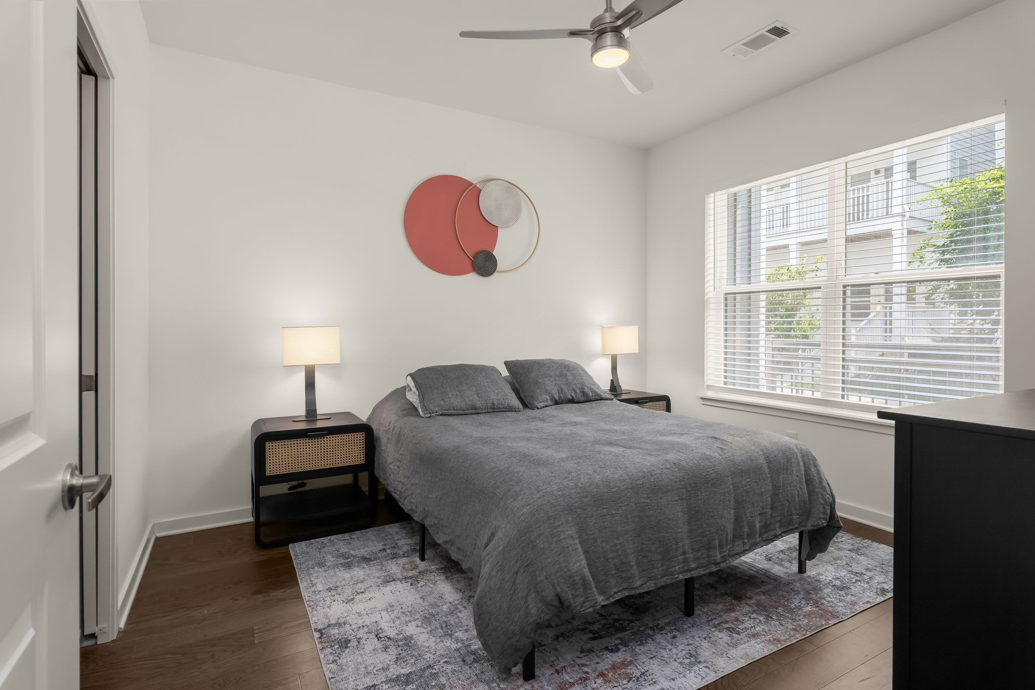 Furnished bedroom with a gray bed, matching nightstands, and a large window with blinds.