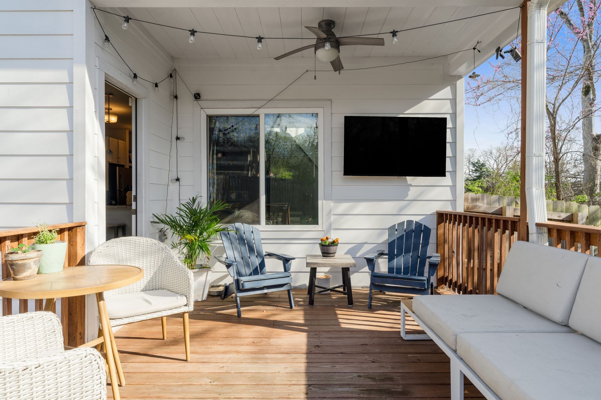 Covered wooden deck with wicker seating, blue Adirondack chairs, string lights, and a wall-mounted television.