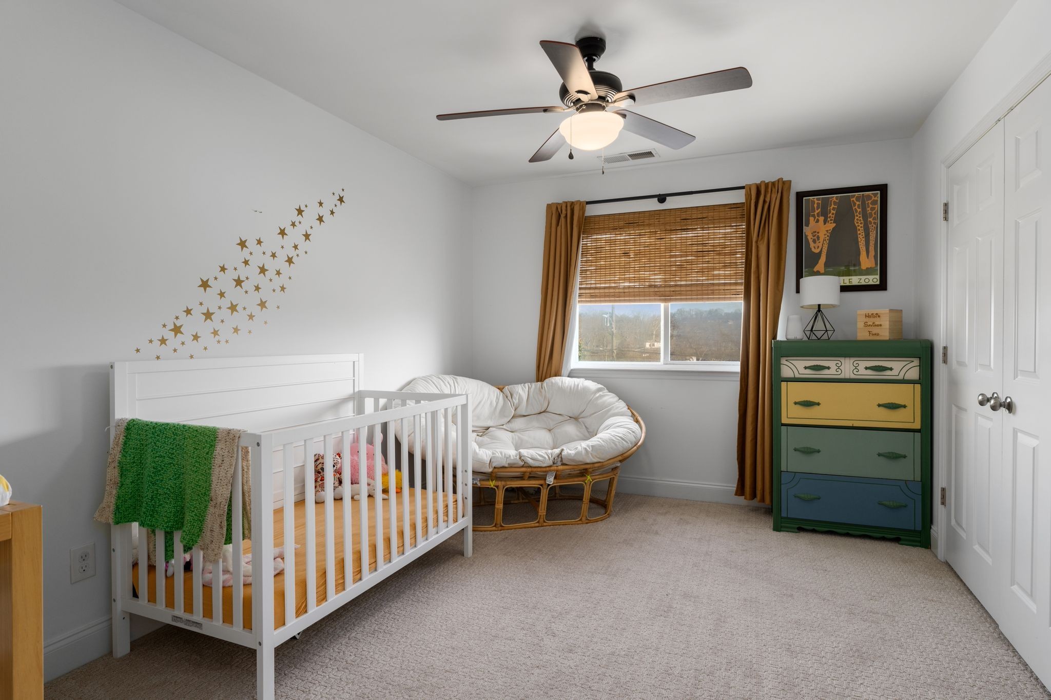 Nursery with a white crib, round cushioned chair, bamboo blinds, and colorful dresser.