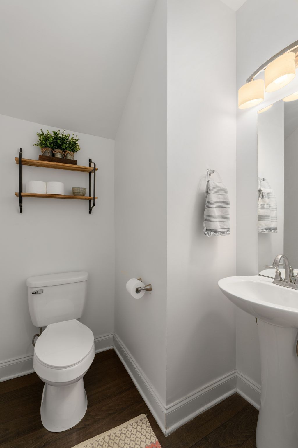 Bathroom with a white toilet, pedestal sink, wooden shelves, and dark flooring.