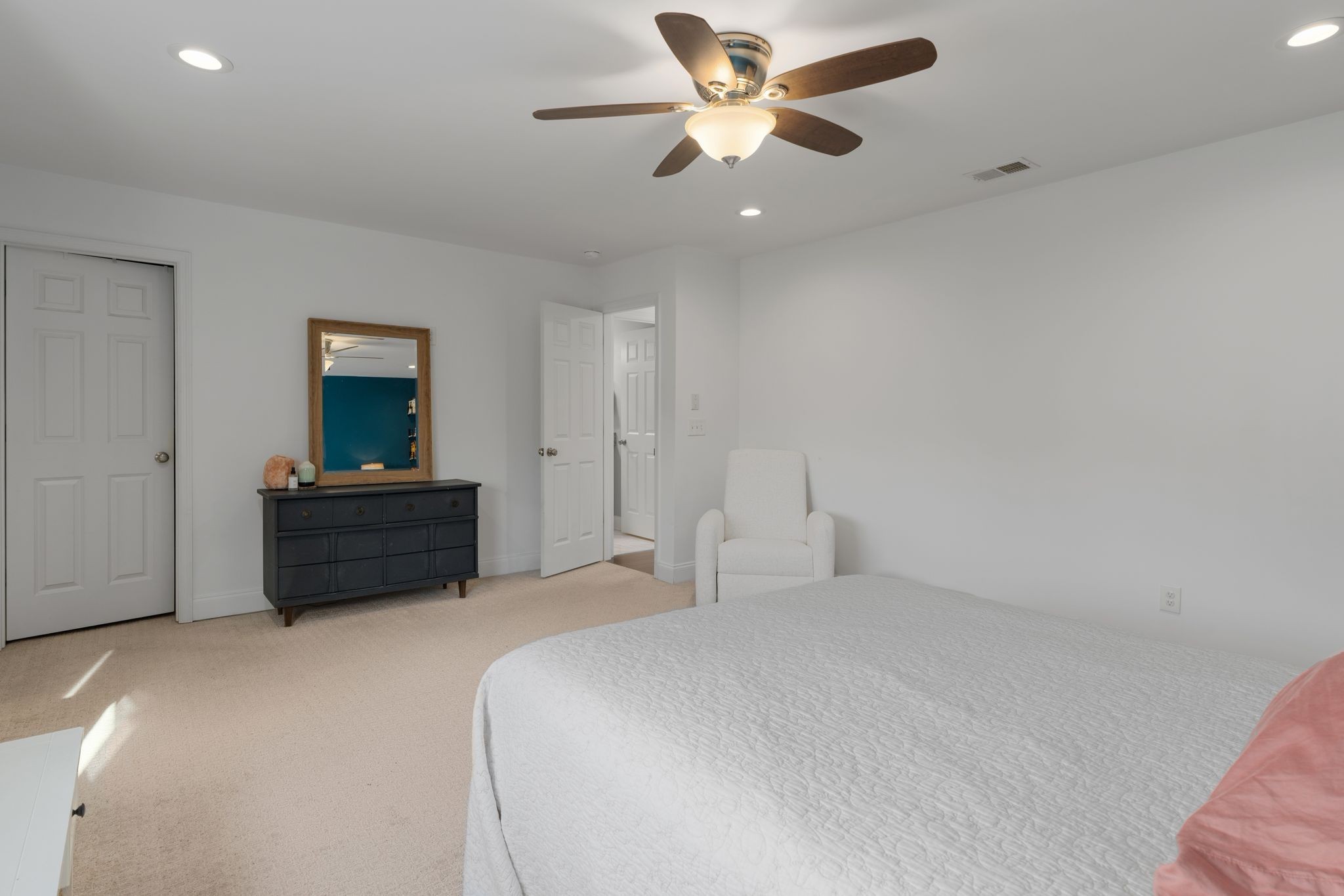 Furnished bedroom with a white bed, ceiling fan, and dark dresser beneath a wood-framed mirror.
