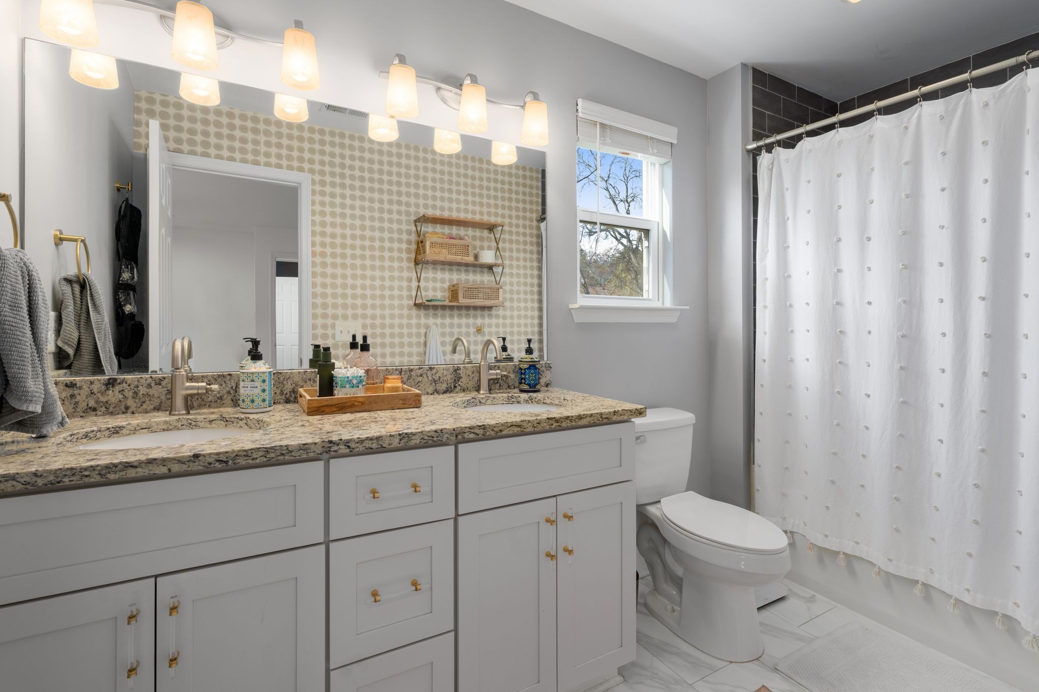 Bathroom with a double-sink vanity, speckled granite counter, patterned wallpaper, and white shower curtain.