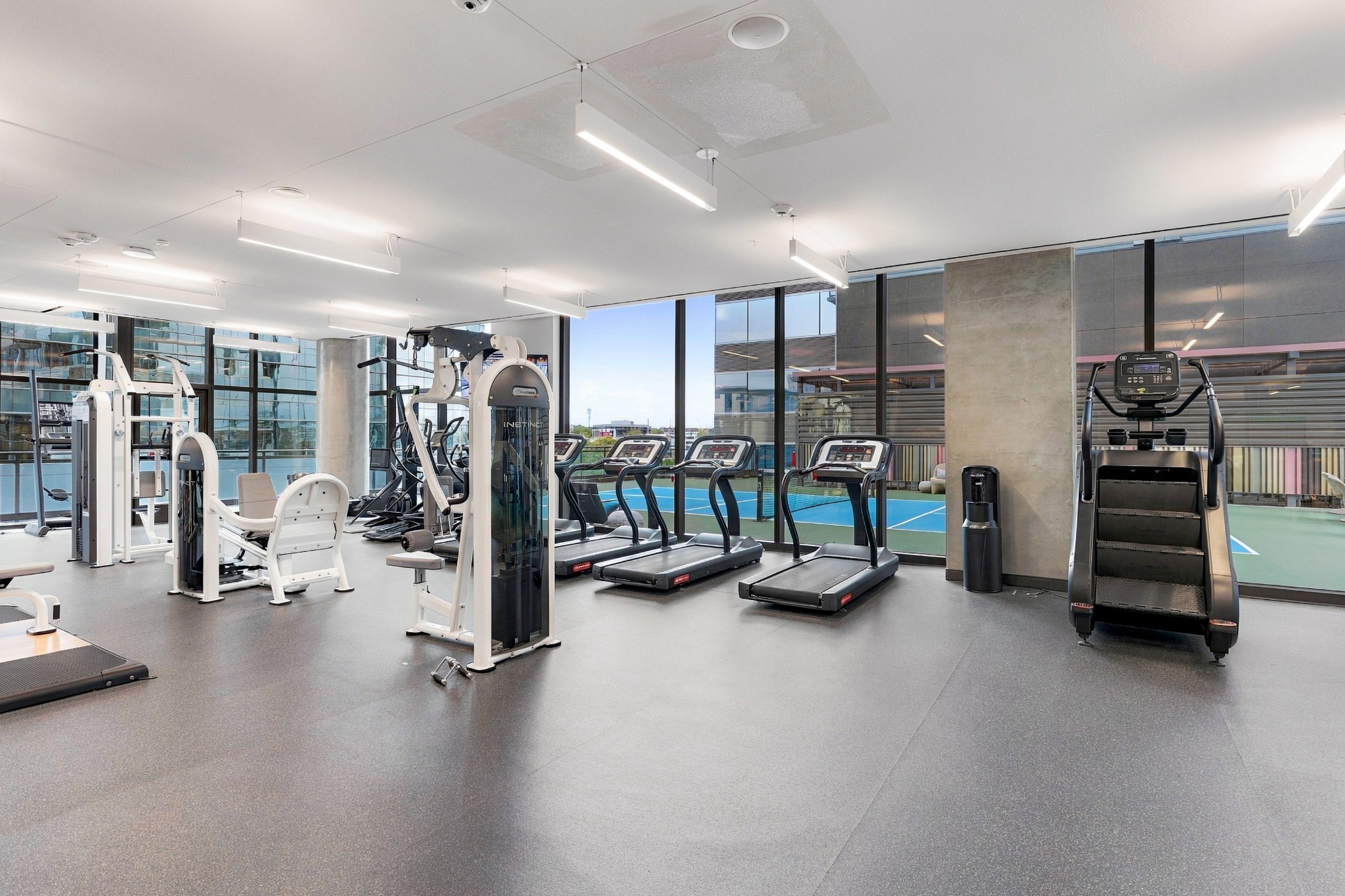 Fitness center with treadmills, strength machines, and large windows overlooking an outdoor court.
