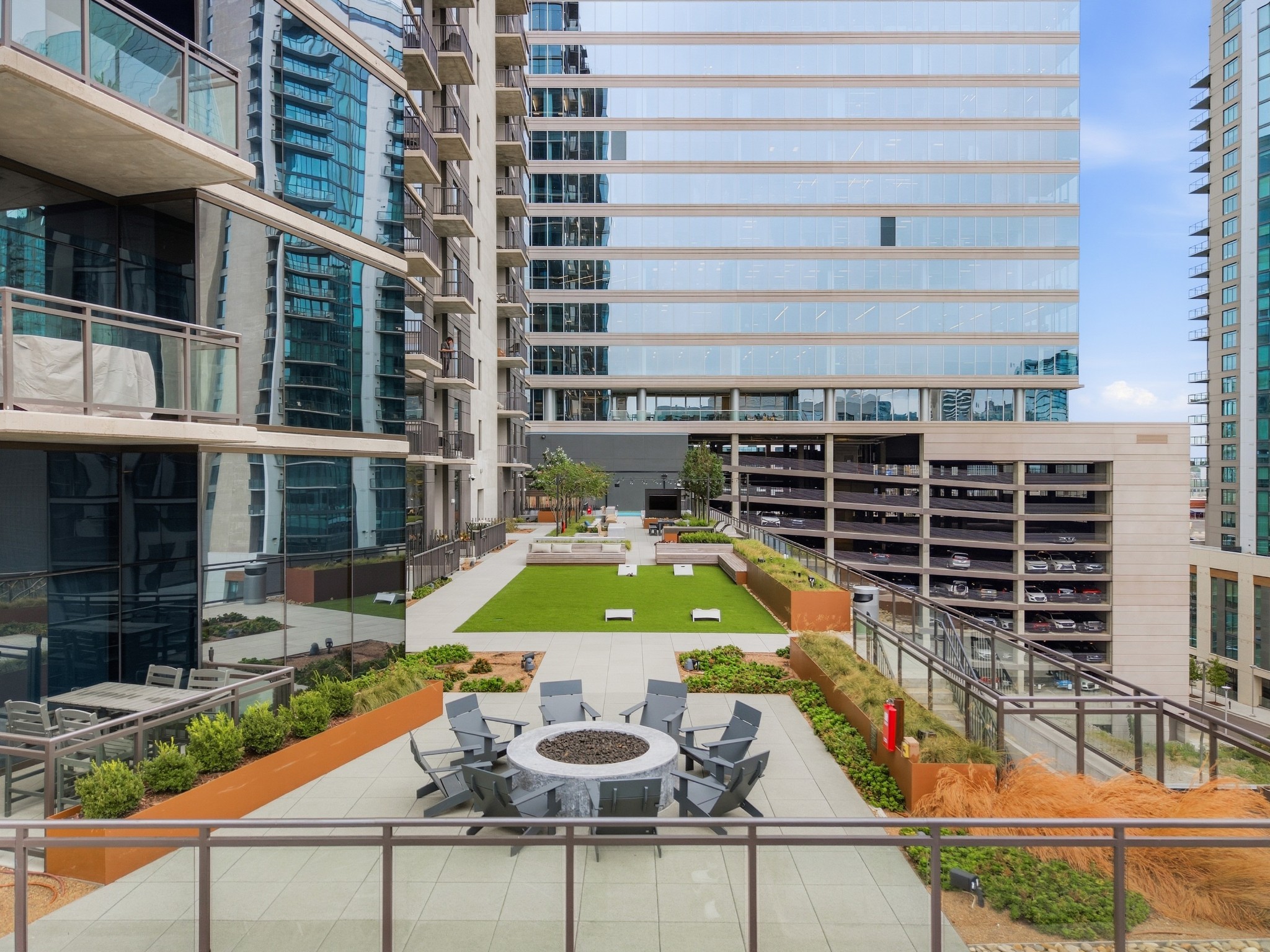 Rooftop terrace with a central fire pit, lounge chairs, and a green turf lawn between high-rises.