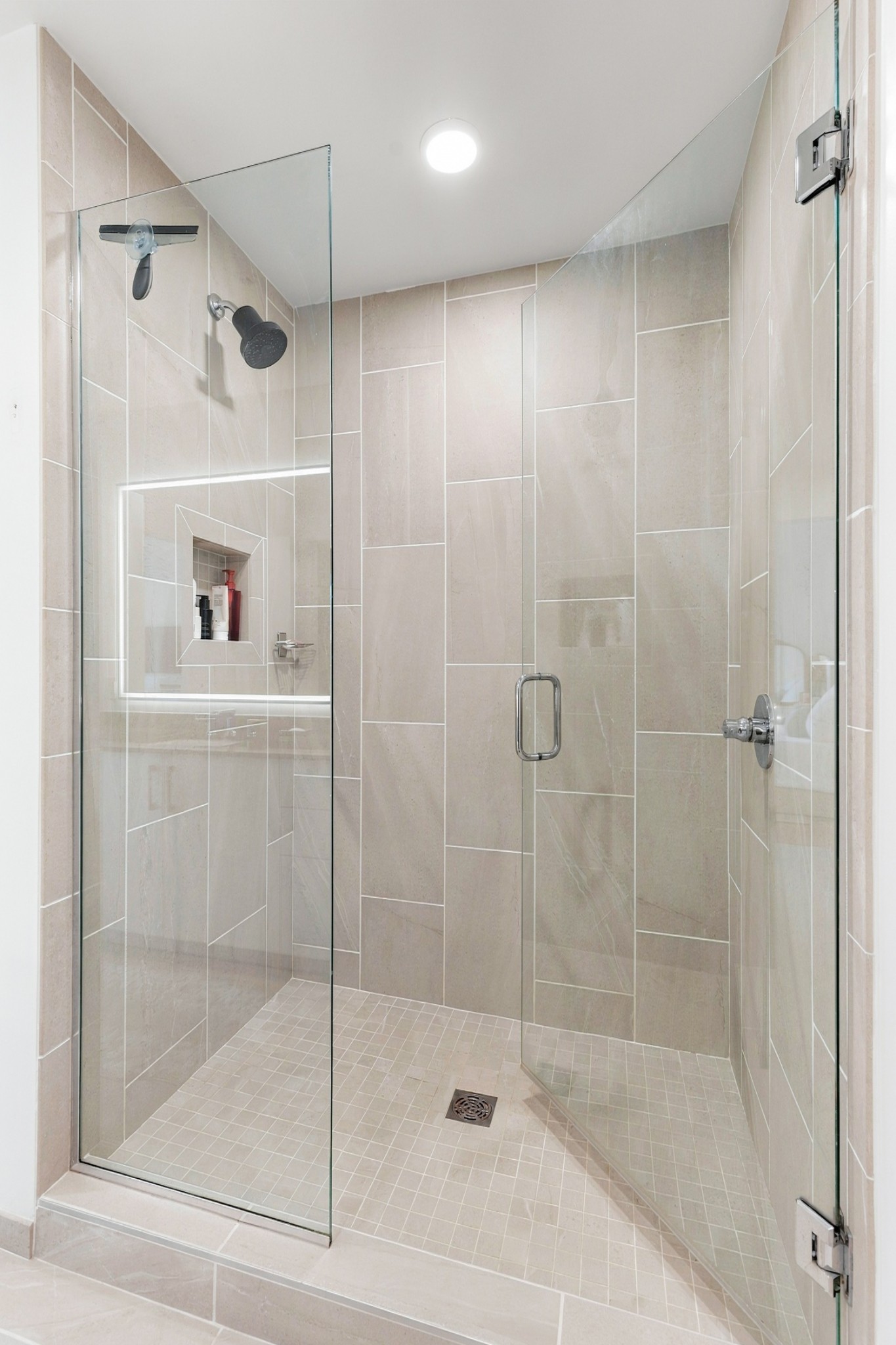Walk-in shower with clear glass doors, beige tile, a built-in niche, and dark fixtures.