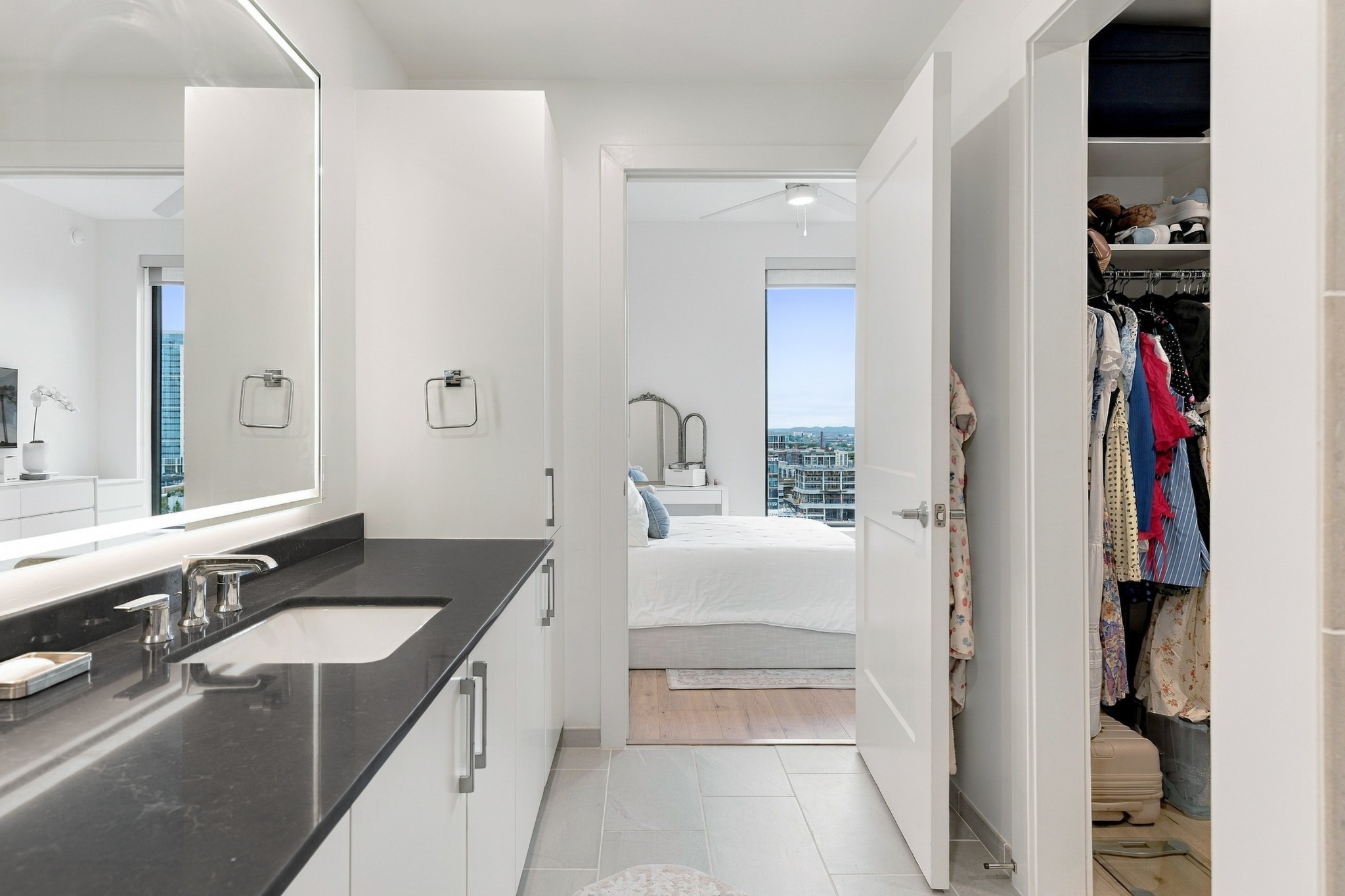 Modern bathroom with a dark vanity opening onto a bedroom and a clothes-filled closet.