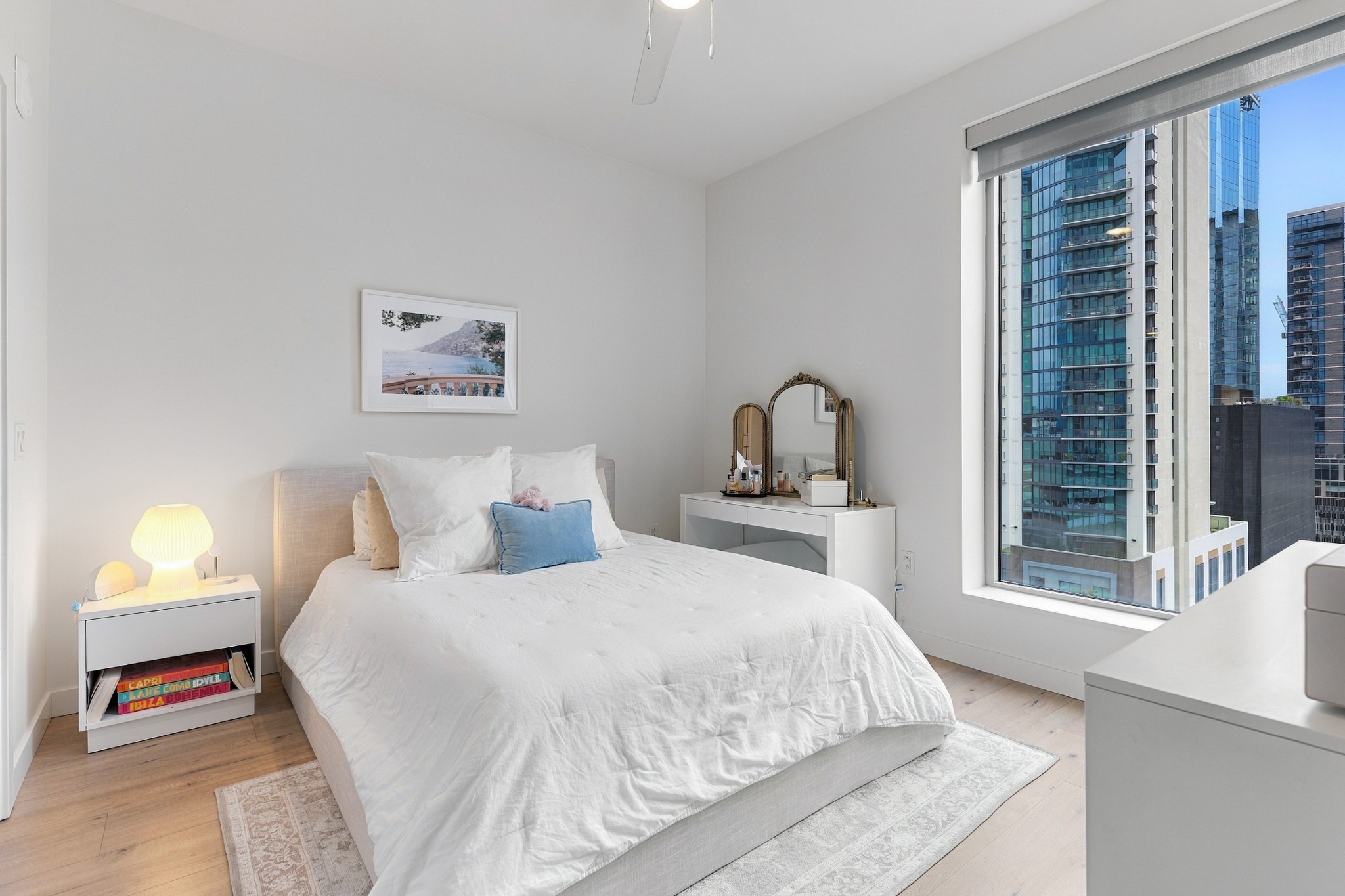 Bright bedroom with a white bed and vanity beside a large window overlooking downtown high-rises.