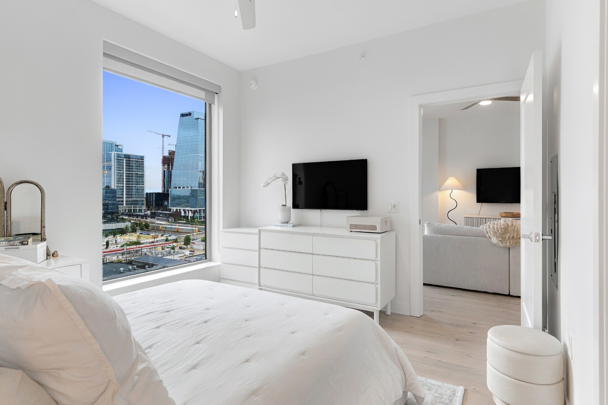 Furnished bedroom with a large window overlooking downtown high-rises and an open doorway to the living area.