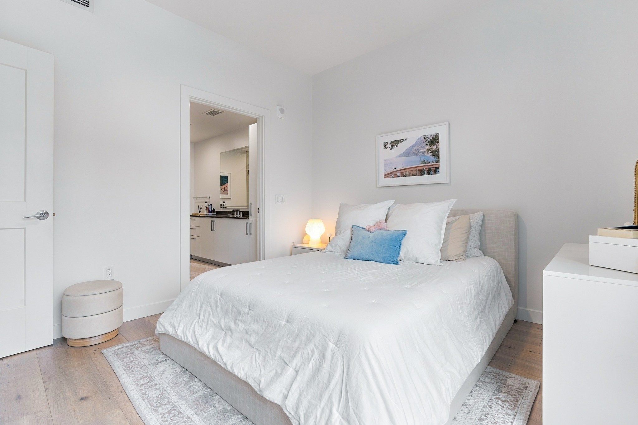 Staged bedroom with a white bed, blue accent pillow, and open doorway to the bathroom.