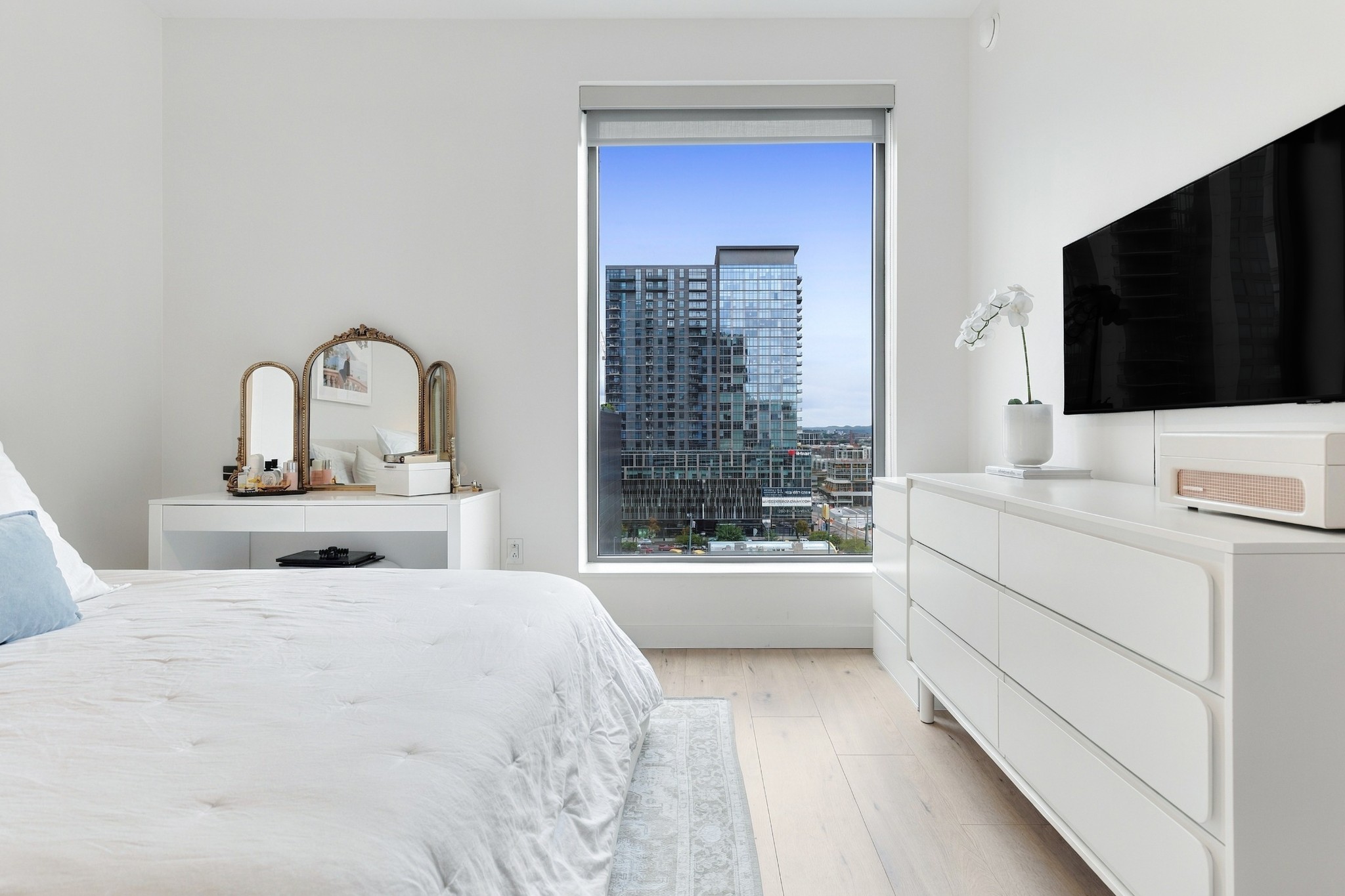 Furnished white bedroom with a large window overlooking a glass high-rise.