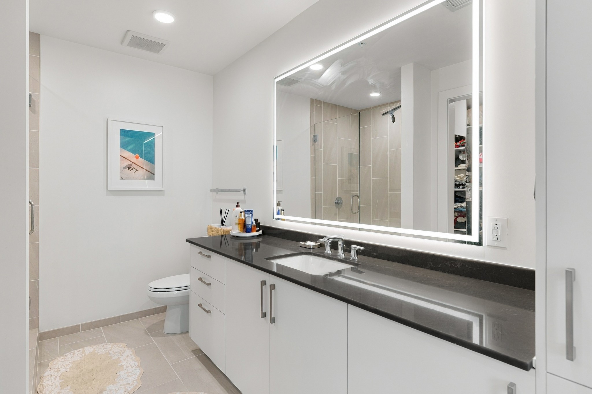 Modern bathroom with a long dark countertop, illuminated mirror, and tiled walk-in shower.