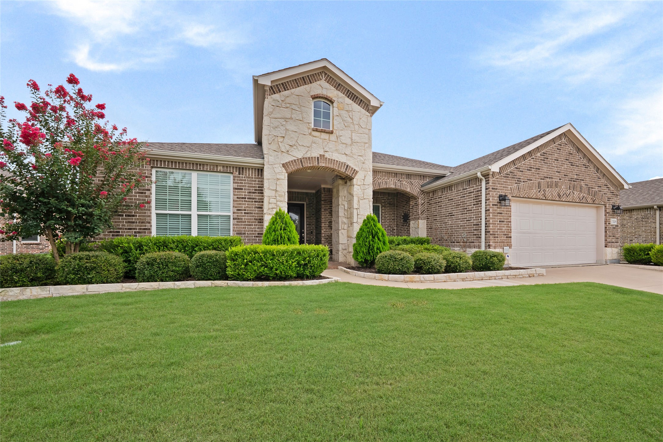 Brick home with a stone entry tower, attached garage, flowering tree, and broad front lawn.