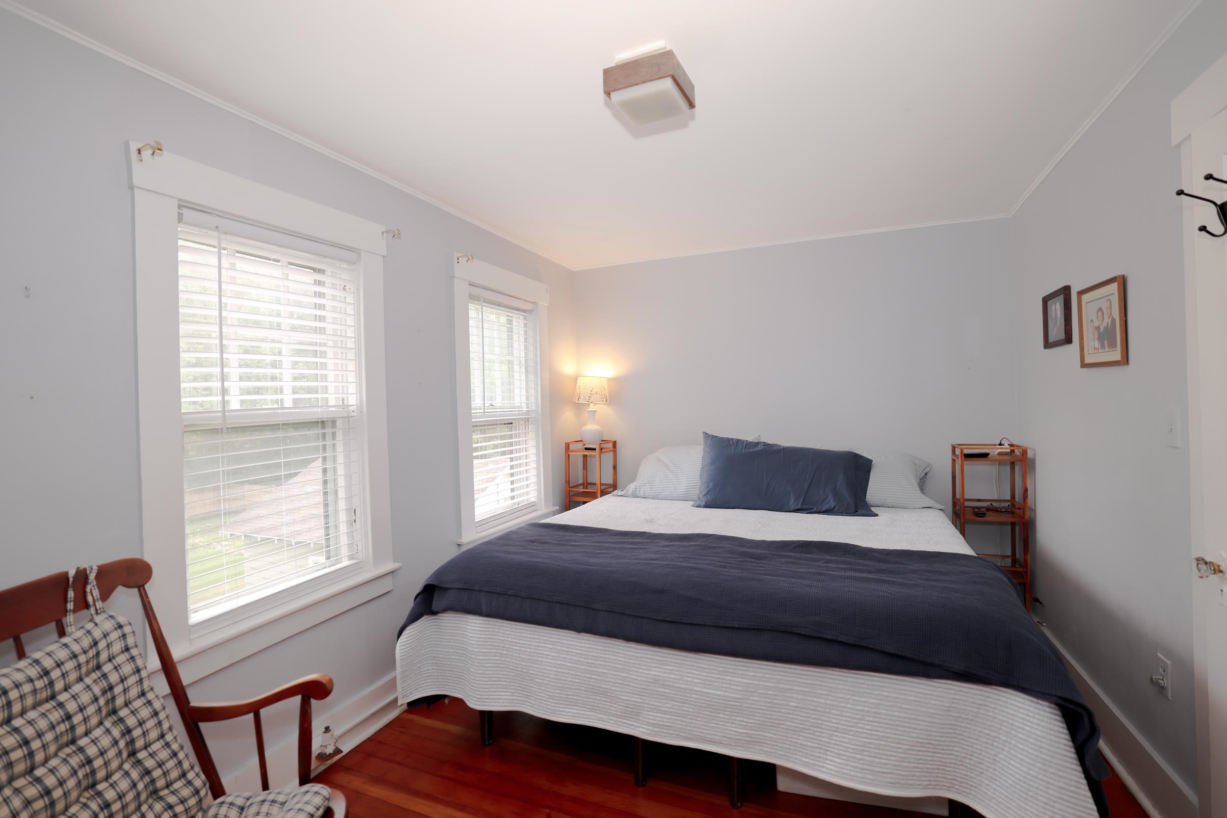 Furnished bedroom with a navy-and-white bed, two blind-covered windows, and reddish hardwood floors.