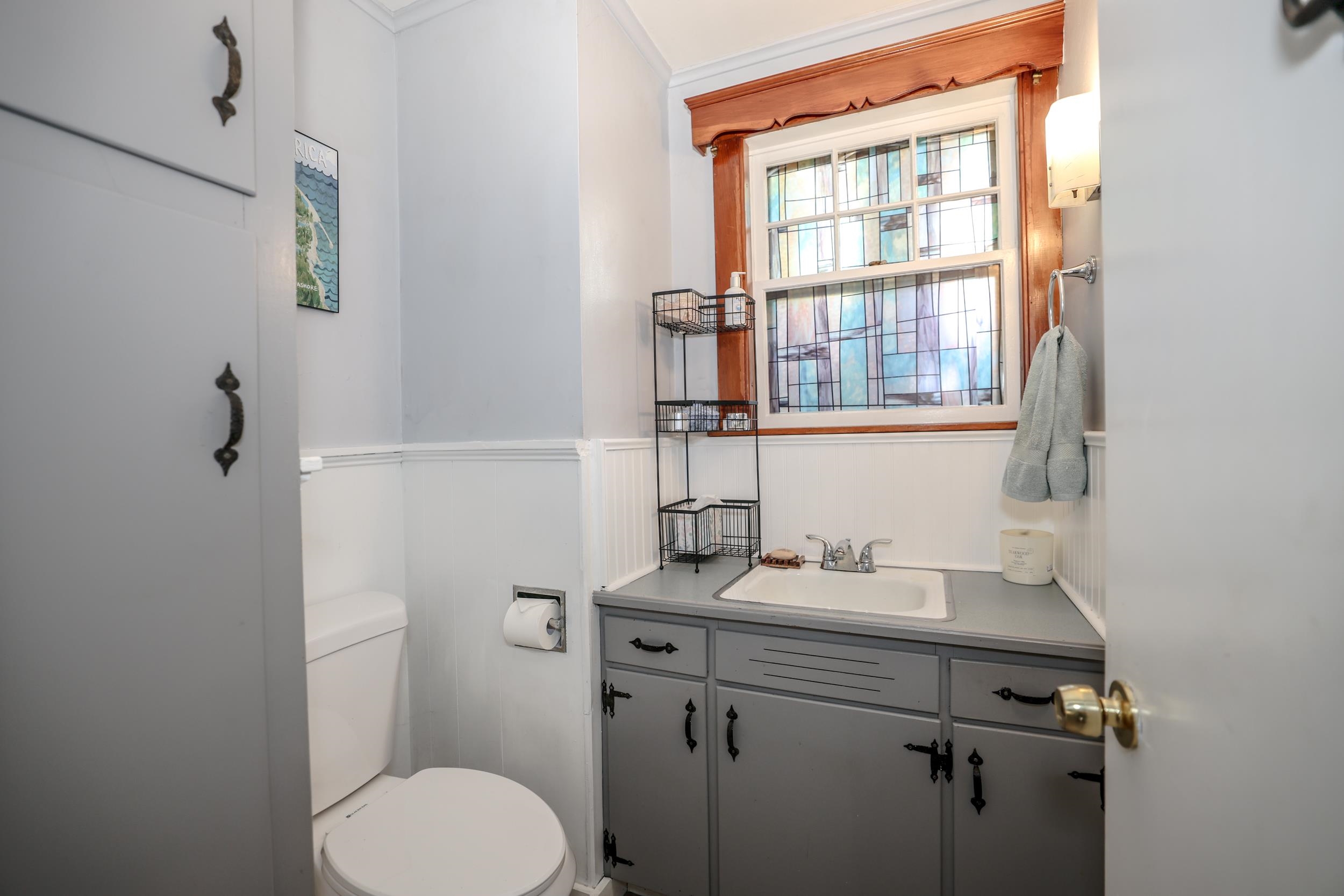 Bathroom with a gray vanity, white beadboard wainscoting, and a large stained-glass window.