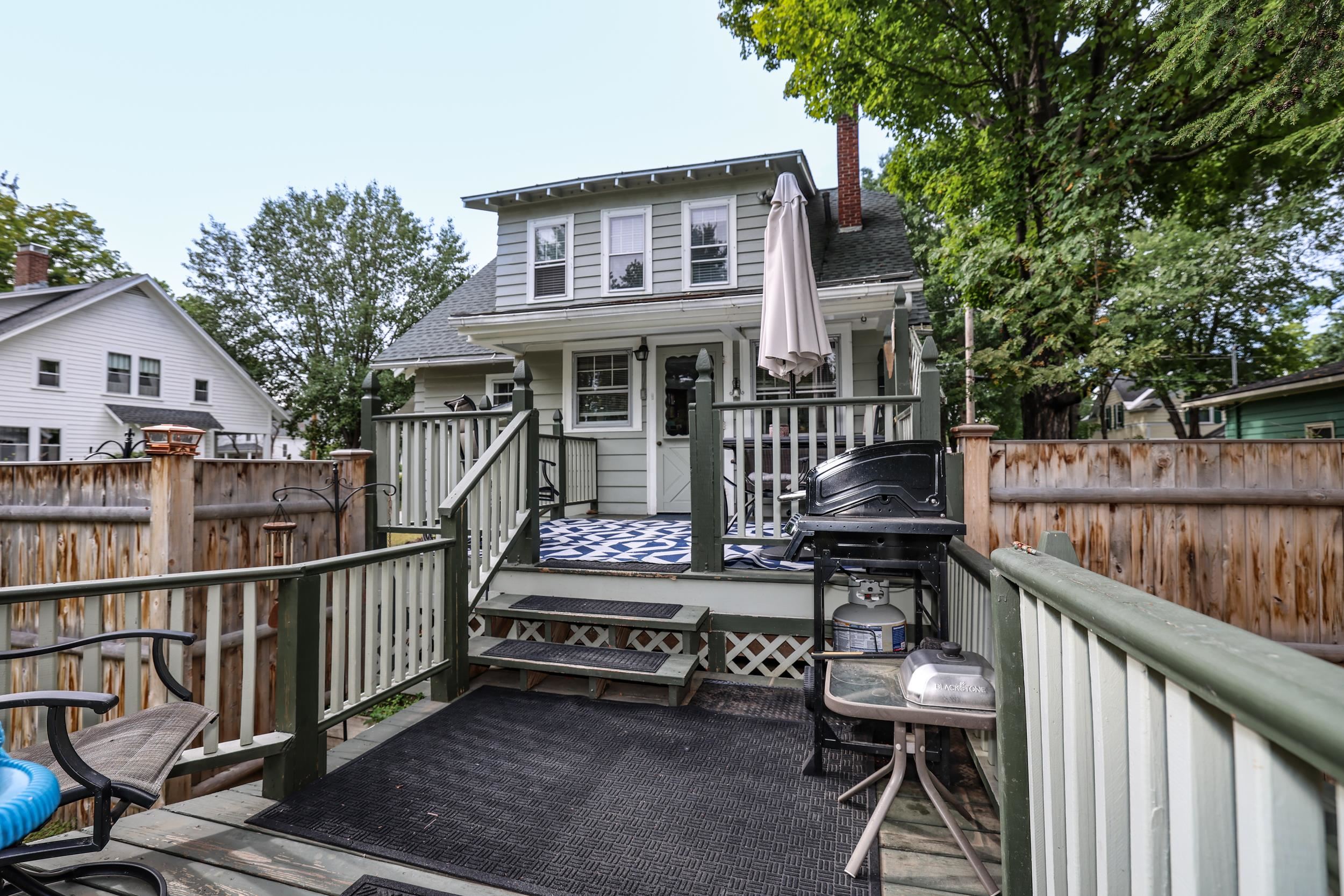Gray two-story house behind a raised deck with green railings, outdoor seating, and a Blackstone griddle.