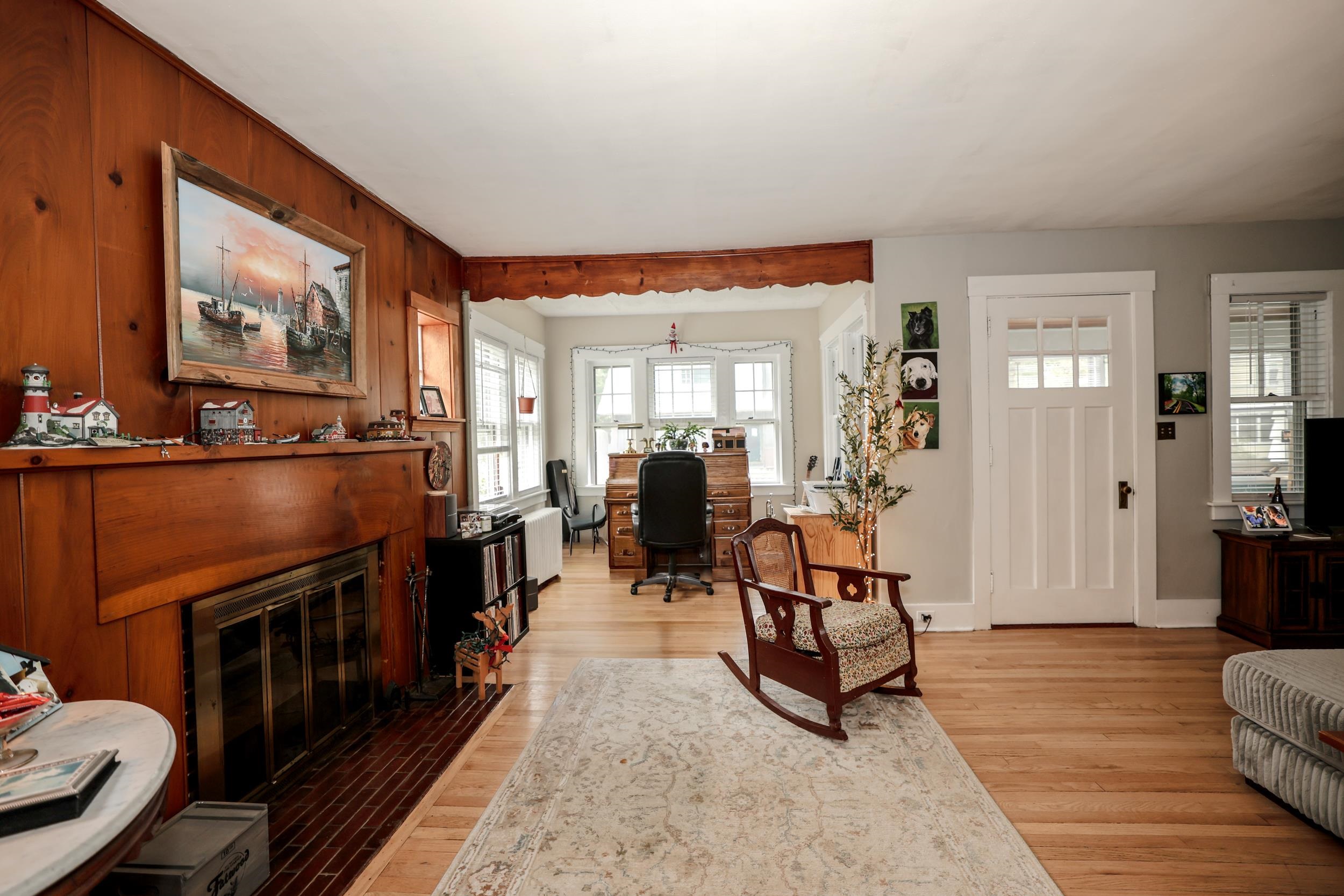 Living room with a wood-paneled fireplace, hardwood floors, and a desk by bay windows.
