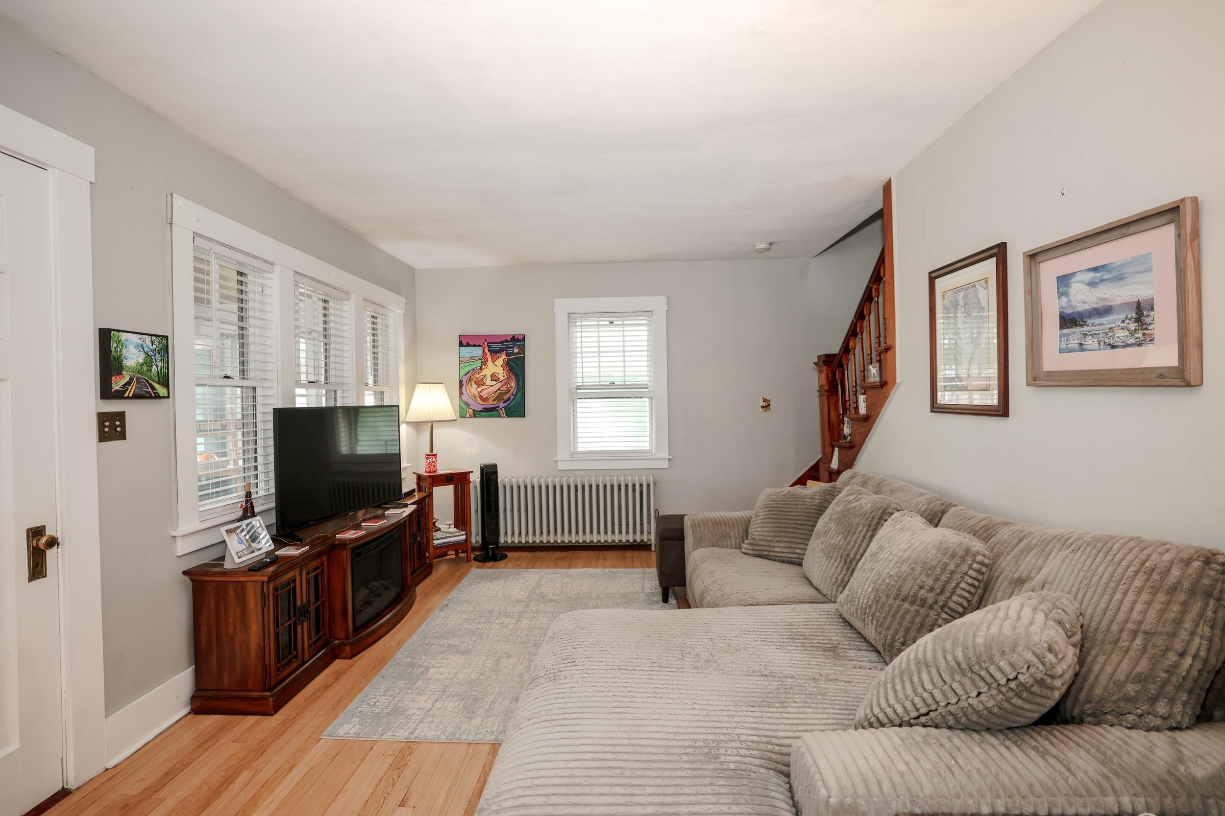 Living room with a gray sectional, hardwood floors, tall windows, and a wooden staircase.