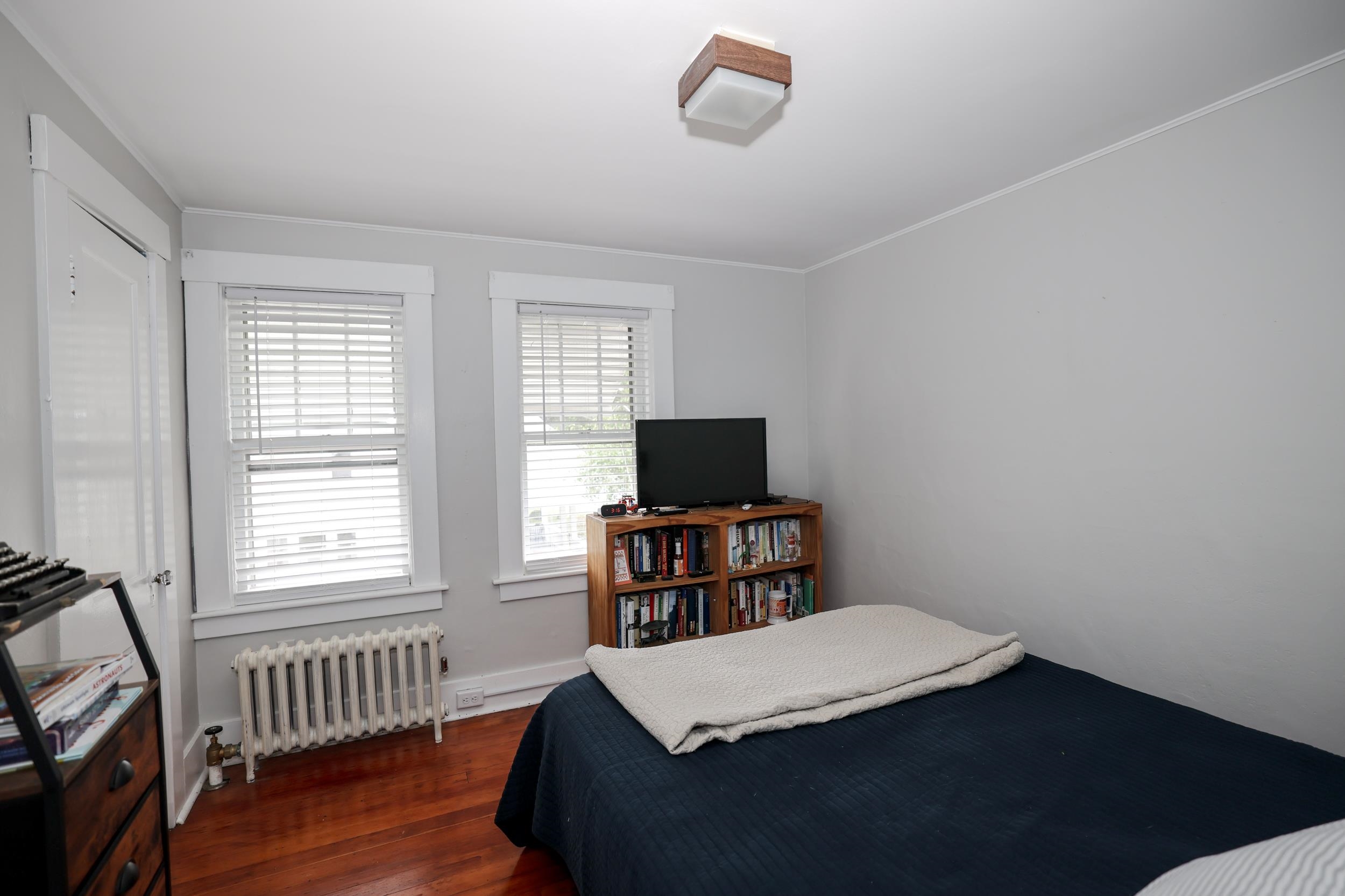 Bedroom with a dark blue bed, hardwood floors, two blind-covered windows, and a bookcase with a television.