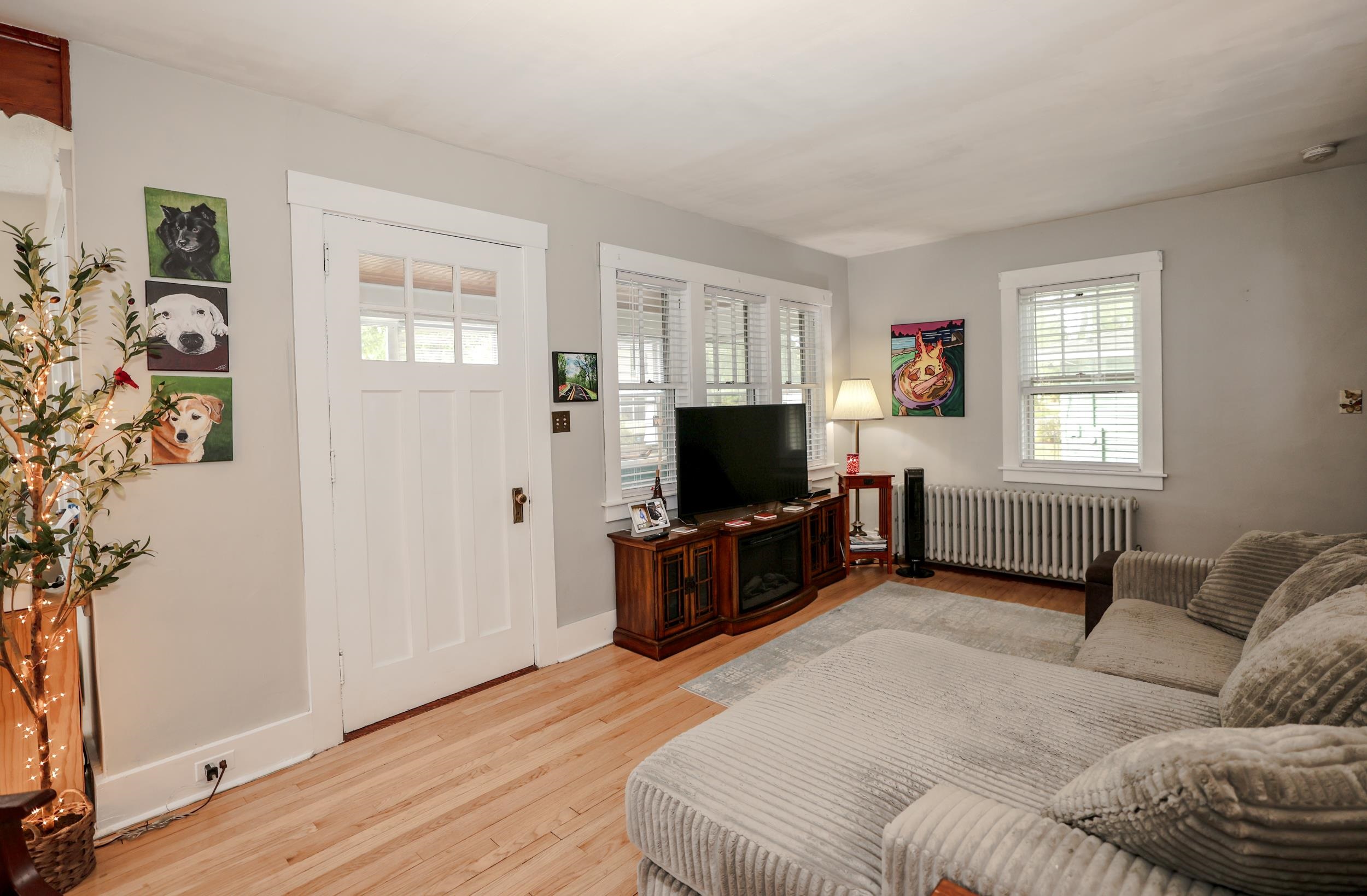 Living room with a gray sectional, hardwood floors, and a television between white-trimmed windows.