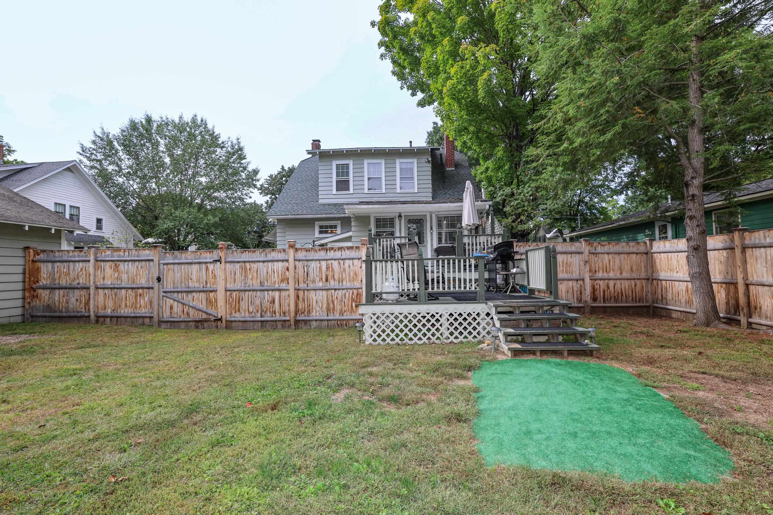 Rear of a gray house with a raised deck and fenced yard beneath mature trees.