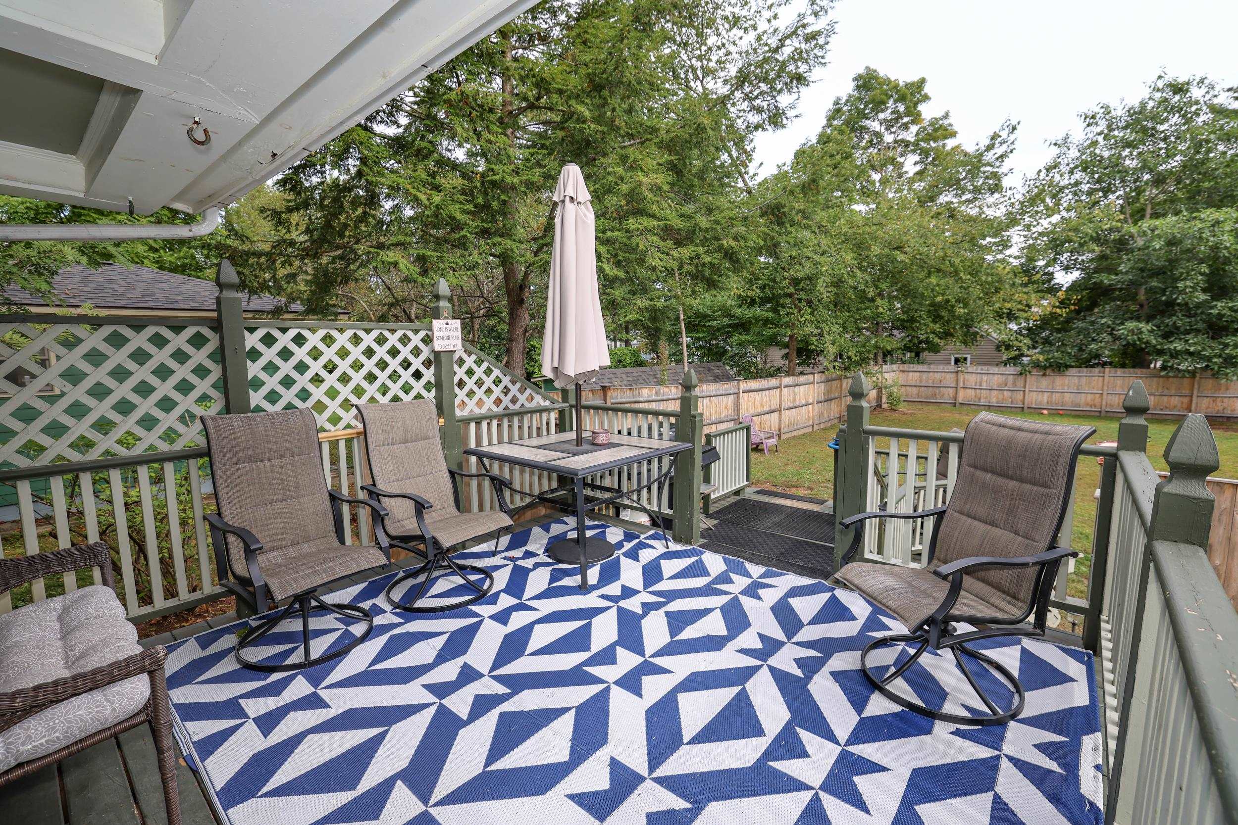 Furnished deck with a geometric blue-and-white rug, patio seating, and a fenced, tree-lined backyard.