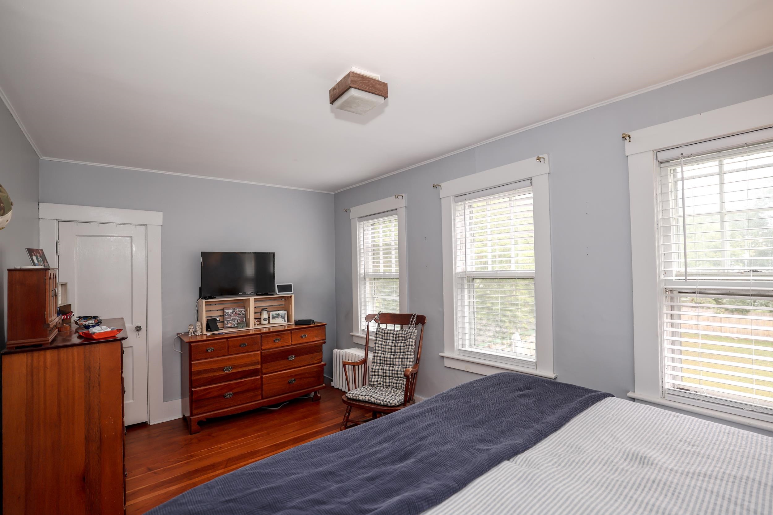 Furnished bedroom with hardwood floors, three bright windows, wooden dressers, and a plaid rocking chair.
