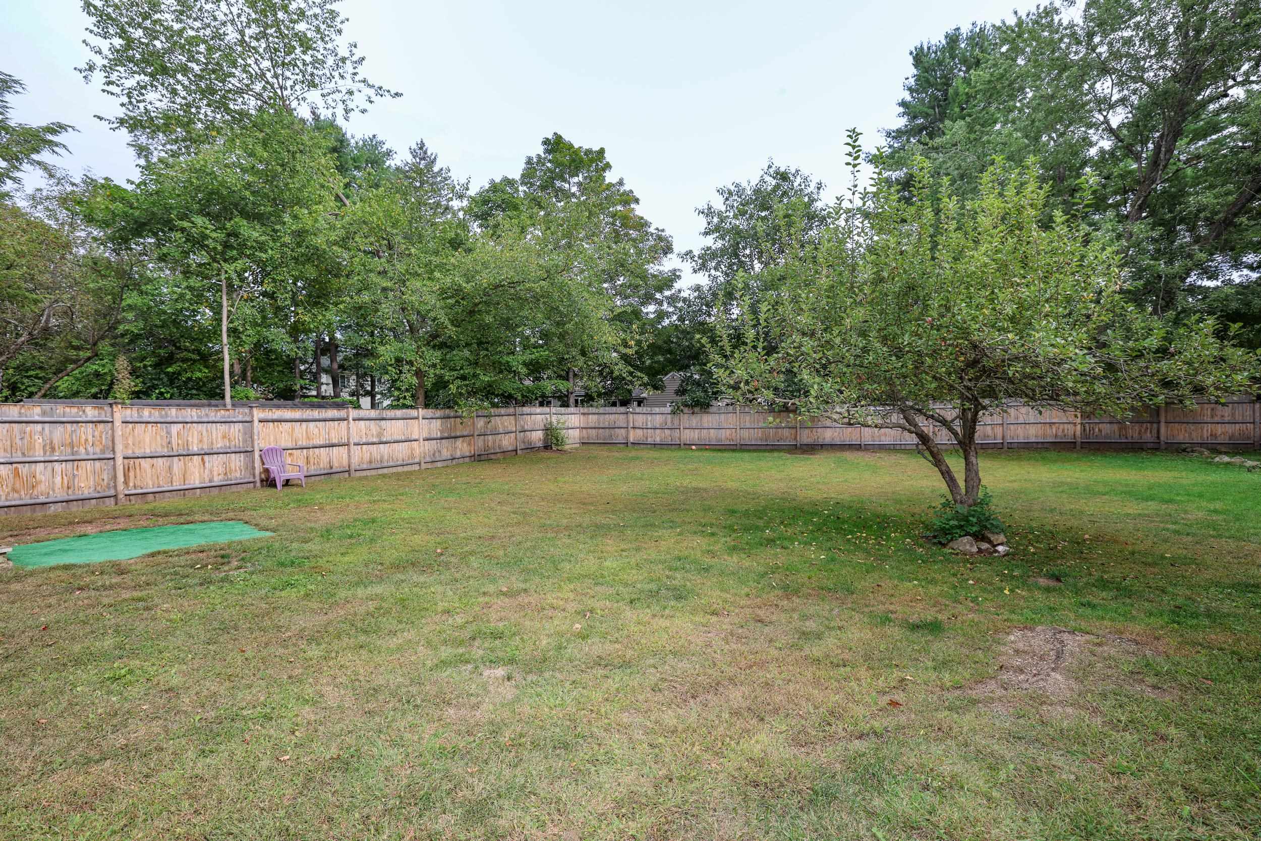 Fenced backyard with a broad lawn, mature trees, and a purple chair beside the wooden fence.